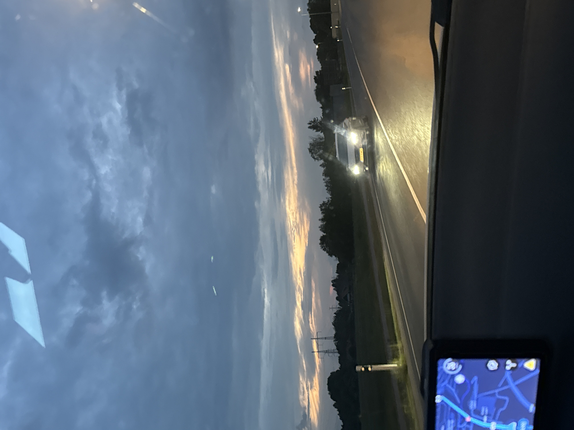 The image captures a forward-facing perspective from inside a vehicle at dusk or early night, on a road identified as being in Saue, Estonia. The sky dominates the upper portion, exhibiting dark blue and grey clouds with a distinct horizontal band of soft orange and pink light along the distant horizon, indicative of a sunset.
In the immediate foreground, the dark-colored dashboard of the recording vehicle is visible. Positioned on the left side of the dashboard is a navigation device, displaying a blue map interface with a bright light-blue route line, various white graphical icons, and a prominent yellow triangular warning symbol. Two light-colored, angular reflections, likely from interior light sources, are visible on the upper part of the windshield.
The two-lane asphalt road extends into the distance, with white dashed lines separating the lanes. A light-colored sedan, possibly white or silver, is approaching in the opposite lane, its headlights fully illuminated and casting a bright glare on the wet road surface. The license plate of this vehicle is present but illegible. The headlights of the recording vehicle illuminate the asphalt directly in front, creating an orange-yellow glow.
To the left of the road, a grassy verge features a white and black reflective marker post. Further in the distance, a dark line of trees and vegetation forms the horizon. Above the treeline, several high-voltage power line towers are clearly visible against the sky. A lone street light is discernible further down the road on the right side. No human figures are explicitly visible, though the presence of the dashboard and navigation device implies a driver within the recording vehicle.
