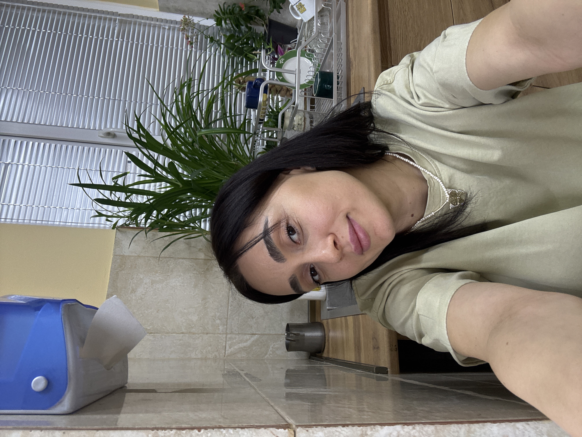 The image displays a female individual taking a selfie in an indoor setting, consistent with a kitchen or utility room. The specific location within Tbilisi, Georgia, cannot be determined from the visual information provided.
The subject, positioned centrally and facing the camera, has dark, shoulder-length hair, dark eyes, dark eyebrows, and pink lips. She is wearing a light olive green short-sleeved t-shirt and a white beaded necklace with a gold-colored butterfly pendant. Her right arm is extended towards the camera, partially visible in the lower right foreground.
The background features light beige/cream square-tiled walls on the left and behind the subject, with a pale yellow wall section above the tiles. A blue and grey wall-mounted paper towel dispenser, with one sheet protruding, is affixed to the upper left wall. A light brown wooden countertop extends across the middle ground. On the left side of the countertop, a dark metallic pitcher or jug is visible. To the right, a metallic sink basin is partially visible, with a small metallic object nearby. A silver-colored metallic dish drying rack, containing a blue cup, a green and white patterned plate, clear glass tumblers, a white mug with a yellow handle, and a small dark green container, rests on the countertop in front of a window. The window is equipped with horizontal white venetian blinds that are largely closed. Several potted green plants, including a prominent one with long, thin leaves and a smaller one with pink flowers, are arranged on the windowsill or countertop in front of the blinds. The scene is uniformly lit.
