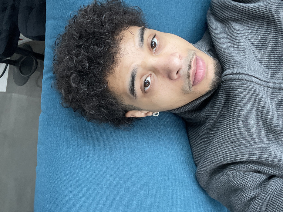 The image, captured in Montigny-le-Bretonneux, France, presents an adult male from an overhead perspective, positioned supine with his head resting on a textured, teal-blue surface. The subject exhibits dark, curly hair with visible gray strands, a neatly trimmed moustache and goatee, and dark brown eyes directed towards the camera. He wears a charcoal gray, ribbed, high-neck sweater featuring a front zipper. A small, circular, possibly jeweled earring is discernible in his left earlobe. The upper right portion of the background, out of focus, reveals components of an indoor environment, including a dark object with a circular base and a section of dark upholstered furniture against a lighter wall.