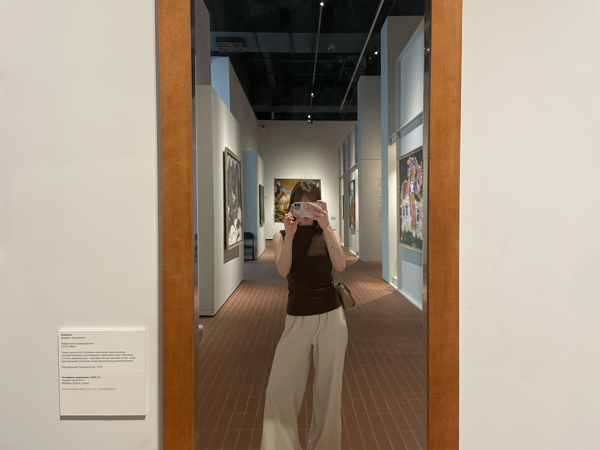 A person is taking a photo of themself in a mirror, which is located in the Perm Art Gallery in Perm, Russia. The person is wearing a brown sleeveless top and white pants. The mirror is framed with a wooden border and is mounted on a white wall next to a sign in Russian describing the mirror. The mirror reflects the art gallery, including paintings on the walls and the brick floor.