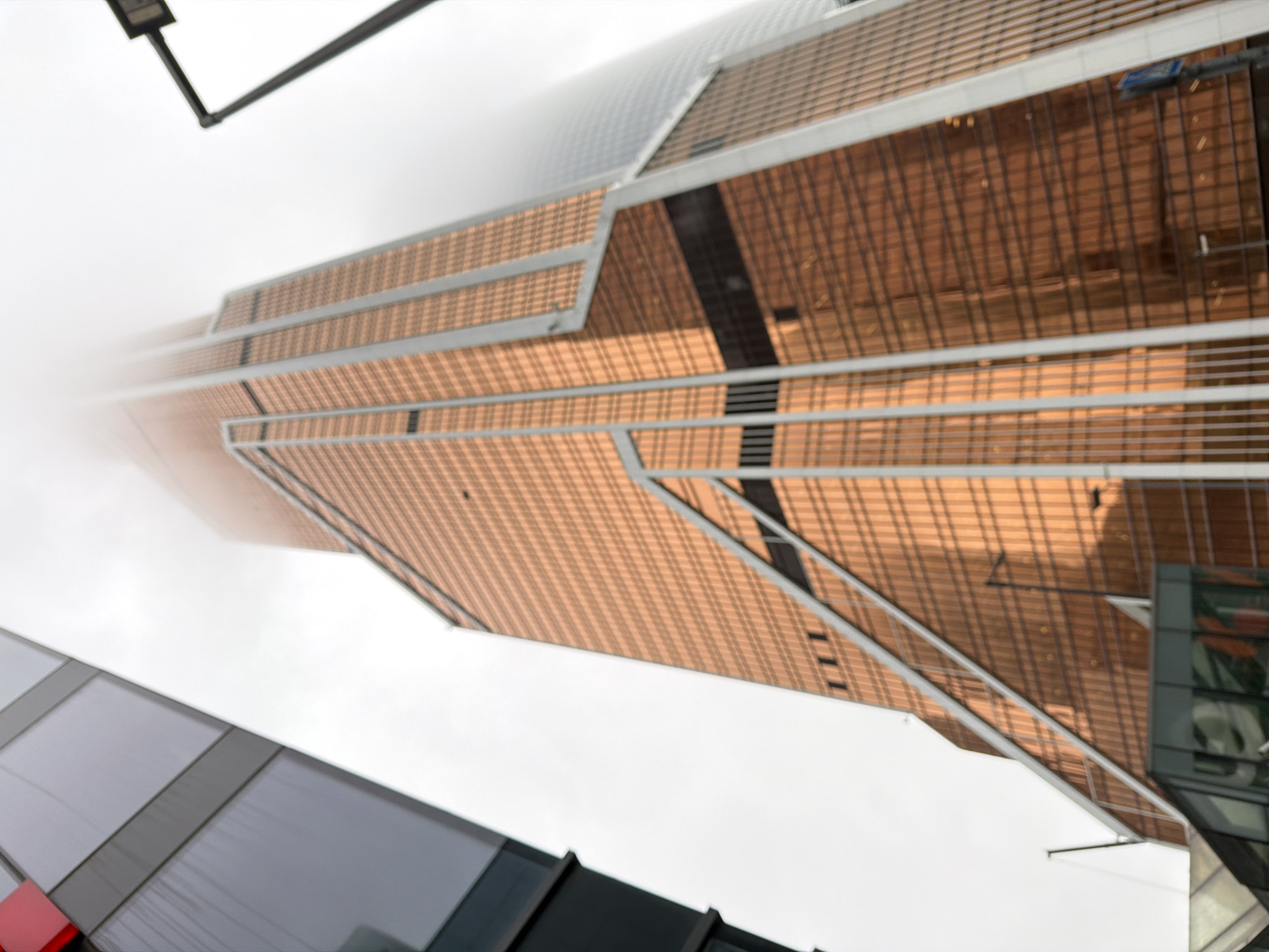 The image shows a view looking up at a modern skyscraper in Moscow, Russia. The building has a distinctive bronze-colored facade with a grid-like pattern of windows. The top of the skyscraper is obscured by clouds or fog, giving the impression of great height. A portion of another building with gray and black panels is visible in the foreground. A street lamp pole is seen extending into the sky. The overall perspective is tilted, creating a dynamic composition.