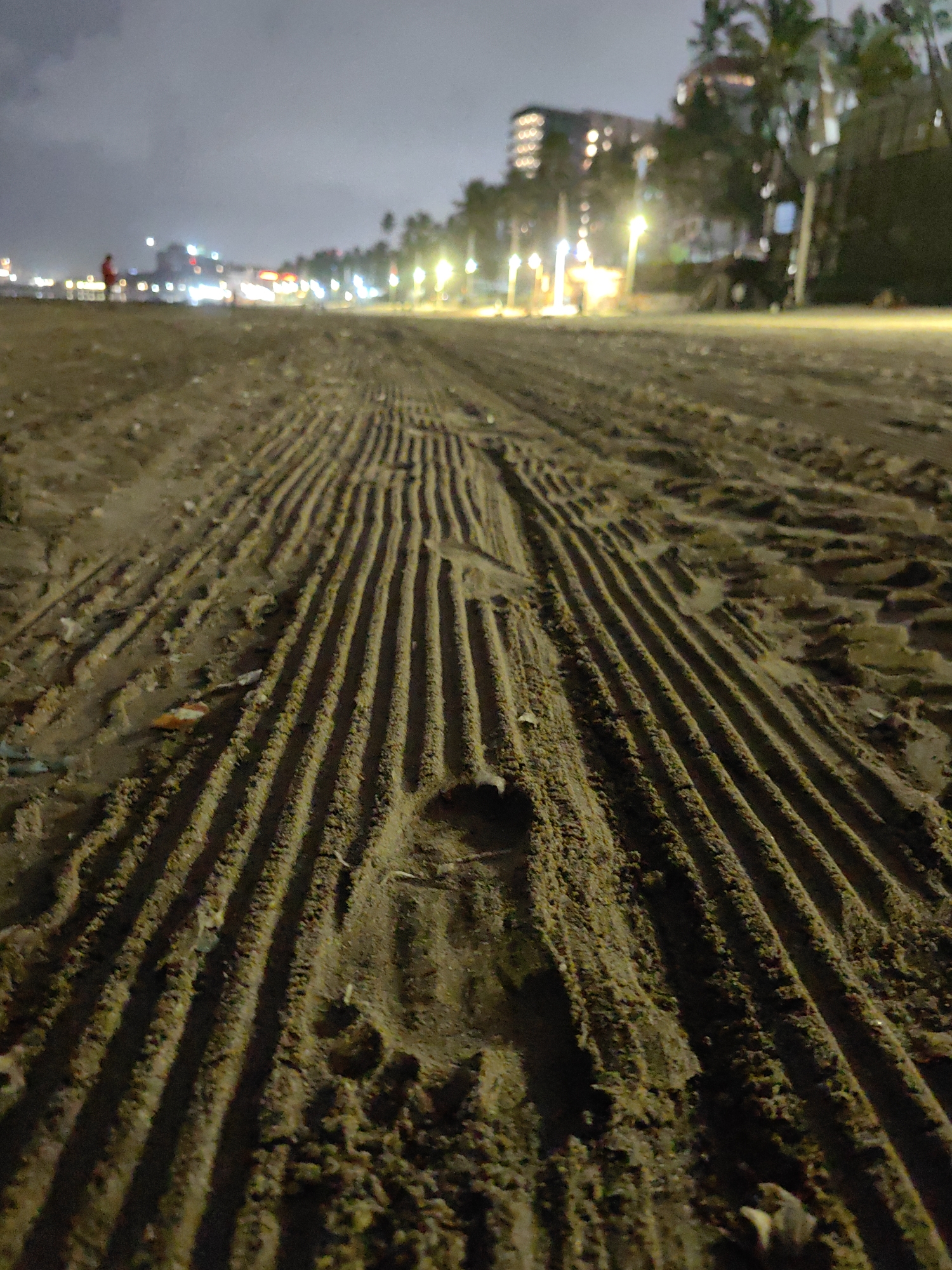 The image is a low-angle shot of a sandy beach at night. The sand is marked with parallel grooves, likely from a vehicle or maintenance equipment. In the foreground, a single footprint is visible in the sand. In the background, a blurred cityscape with illuminated buildings and palm trees is visible. A single figure, possibly a person, is standing on the beach in the distance, facing the water. The sky is dark and cloudy. The scene appears to be located on a beach in Mumbai, India, possibly Juhu Beach or Chowpatty Beach, given the presence of a beach at night with city lights and palm trees.