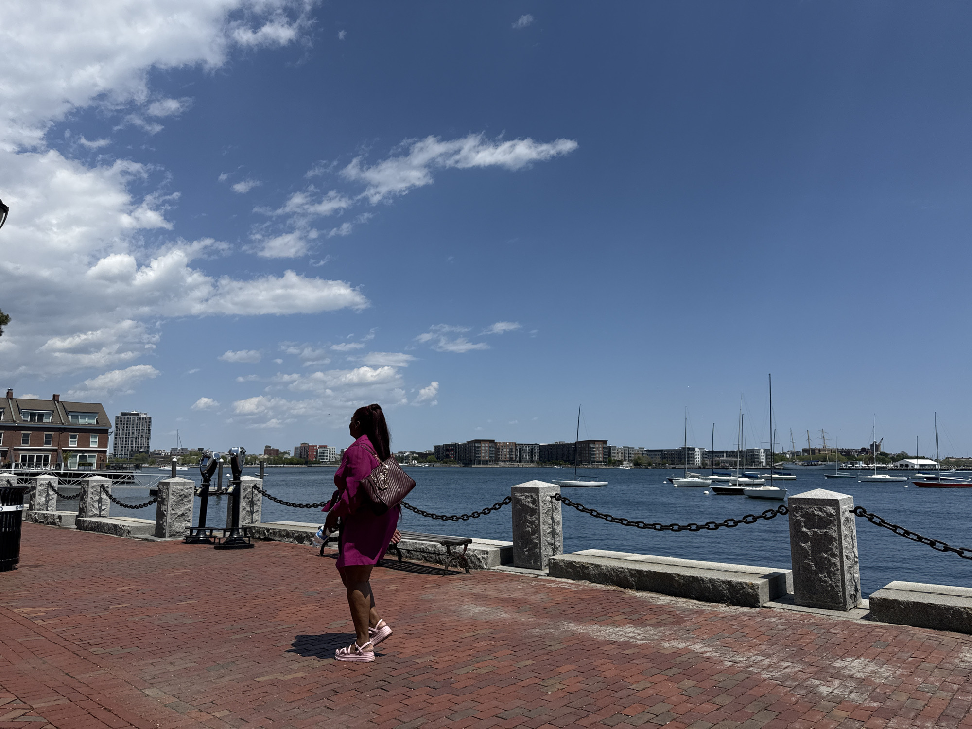 The image depicts a woman in a pink dress and shoes walking along a brick walkway by the water in Boston, Massachusetts. She is carrying a purse and a water bottle. The walkway is bordered by a stone wall with a chain link fence. The Boston harbor is visible, with sailboats docked on the water. Buildings are visible in the background and the sky is blue with fluffy white clouds. There are coin operated tourist binoculars near a trash receptacle on the walkway.