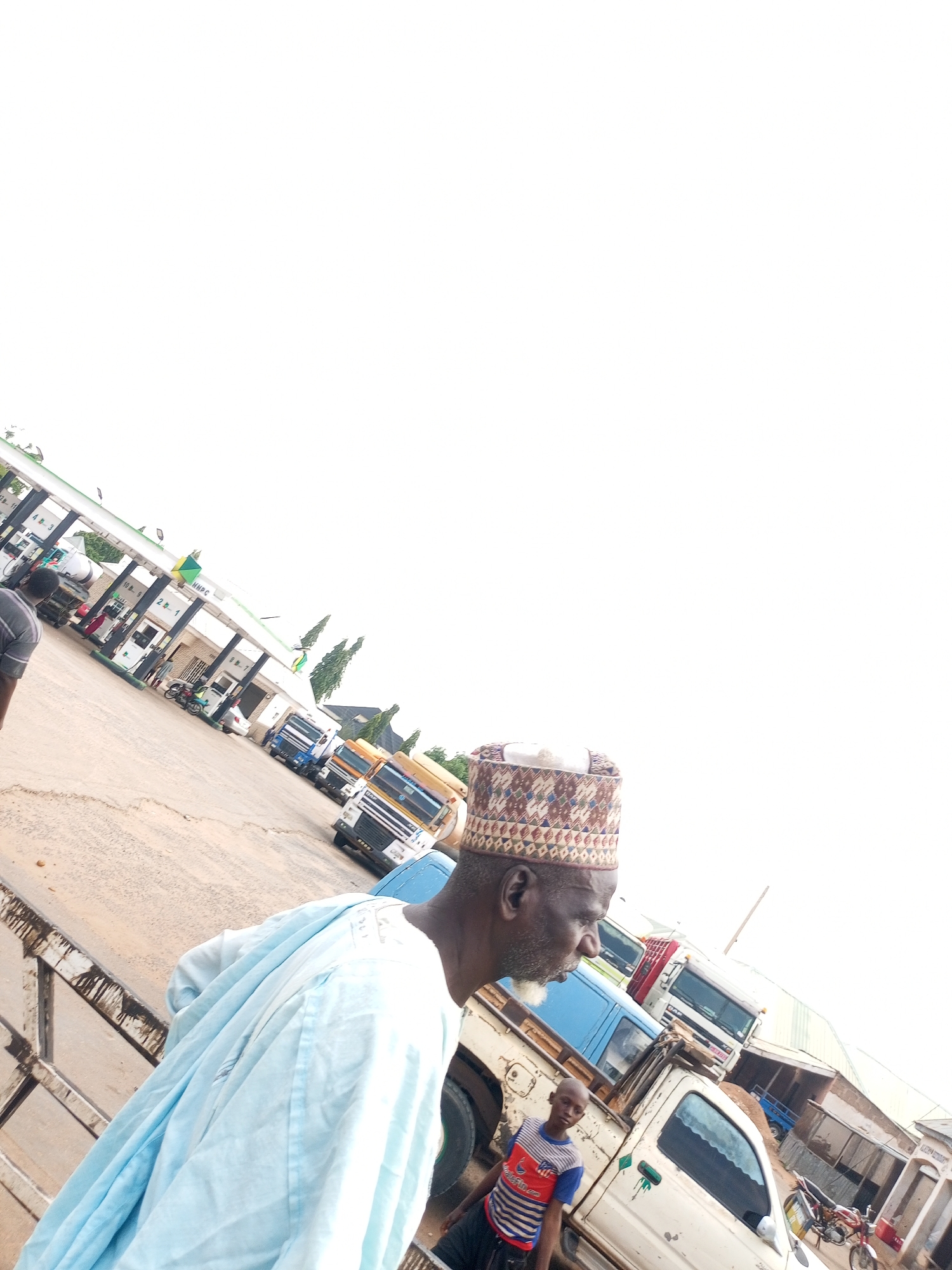 The image appears to be taken at a petrol station in Tempure, Nigeria. An older man in traditional attire, specifically a patterned cap, is partially visible in the foreground on the left. A young boy in a striped shirt stands near a white pick-up truck. Several large trucks are parked in the station area. A motorcycle is parked nearby. The petrol station features green and white canopies with gas pumps underneath. The sky is overcast.