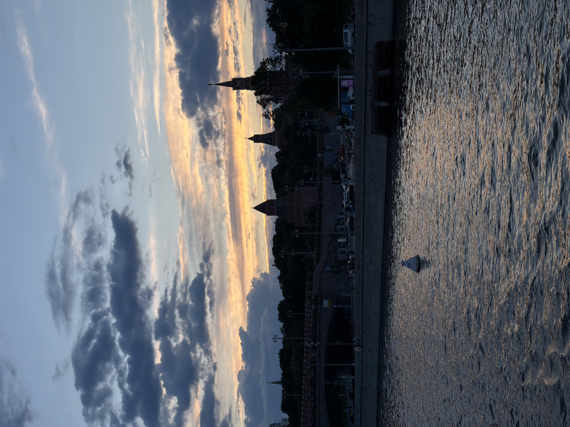 This image captures a scenic view of a city at dusk or sunset, overlooking a river. The setting sun casts a warm, golden glow across the sky and the water, creating a serene atmosphere.
In the upper portion of the frame, the sky transitions from a light blue to a vivid orange and yellow where the sun's light is most intense, indicating the late hour. Various clouds, ranging from wispy white streaks to darker, heavier grey formations, are scattered across the sky.
The middle ground features a dark silhouette of a city skyline, strongly suggestive of the Moscow Kremlin with its distinctive towers and fortifications. Several prominent spires and domed structures can be identified against the glowing sky. Trees line the embankment, which also shows signs of urban activity, including streetlights, hints of vehicles, and possibly pedestrians.
The lower part of the image is dominated by the river, likely the Moskva River, with its surface covered in small, shimmering ripples. The water beautifully reflects the golden and orange hues of the sunset. A white buoy, marked with the number "15," floats prominently in the river. The riverbank or quay wall is visible along the left edge of the river, defining the city's edge.
The overall scene is peaceful, highlighting the architectural grandeur of the city against a dramatic sunset sky.