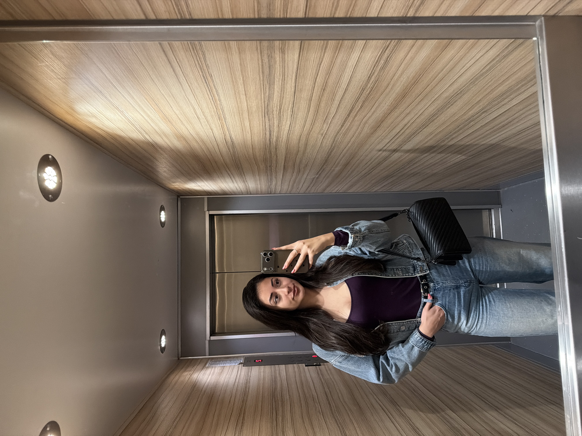 The image shows a woman taking a selfie in an elevator. The elevator has wooden paneling on the ceiling and one wall, with small circular lights embedded in the other wall. The elevator door is visible in the background. The woman is wearing jeans, a dark top, and a denim jacket. She is holding a smartphone in her right hand. She is also wearing a black purse. The location appears to be Sofia, Bulgaria.