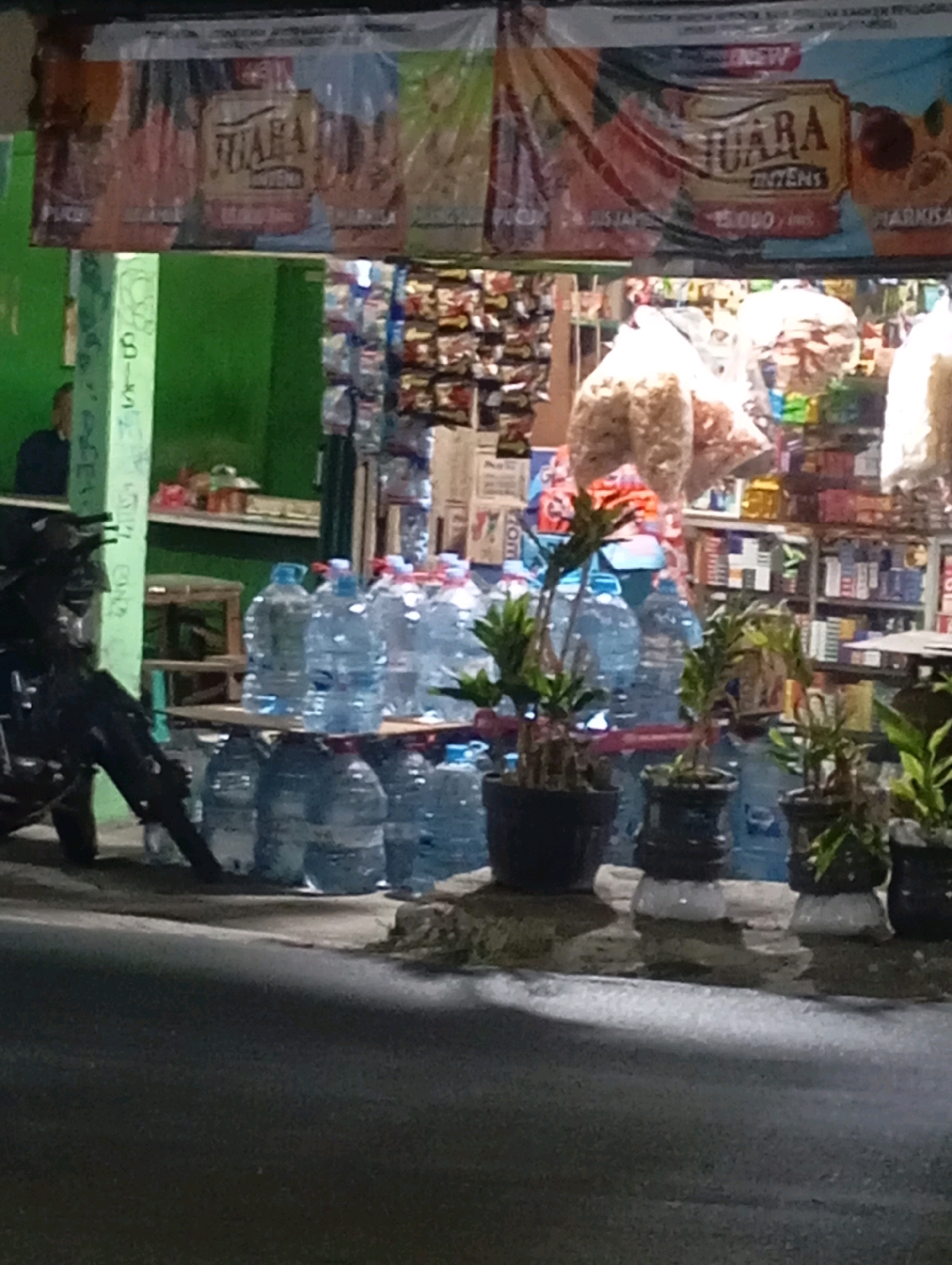 The image depicts a roadside stall in Babelan, Indonesia, at what appears to be dusk or evening. The stall is stocked with bottled water, packaged snacks, and potted plants. A motorcycle is parked on the left side of the frame, partially obstructing the view of the stall. In the background, shelves are filled with various products, suggesting a small convenience store or warung. A banner with text, possibly advertising a product called "JUARA," is hung above the stall. The overall scene is dimly lit, with some artificial lighting casting shadows.