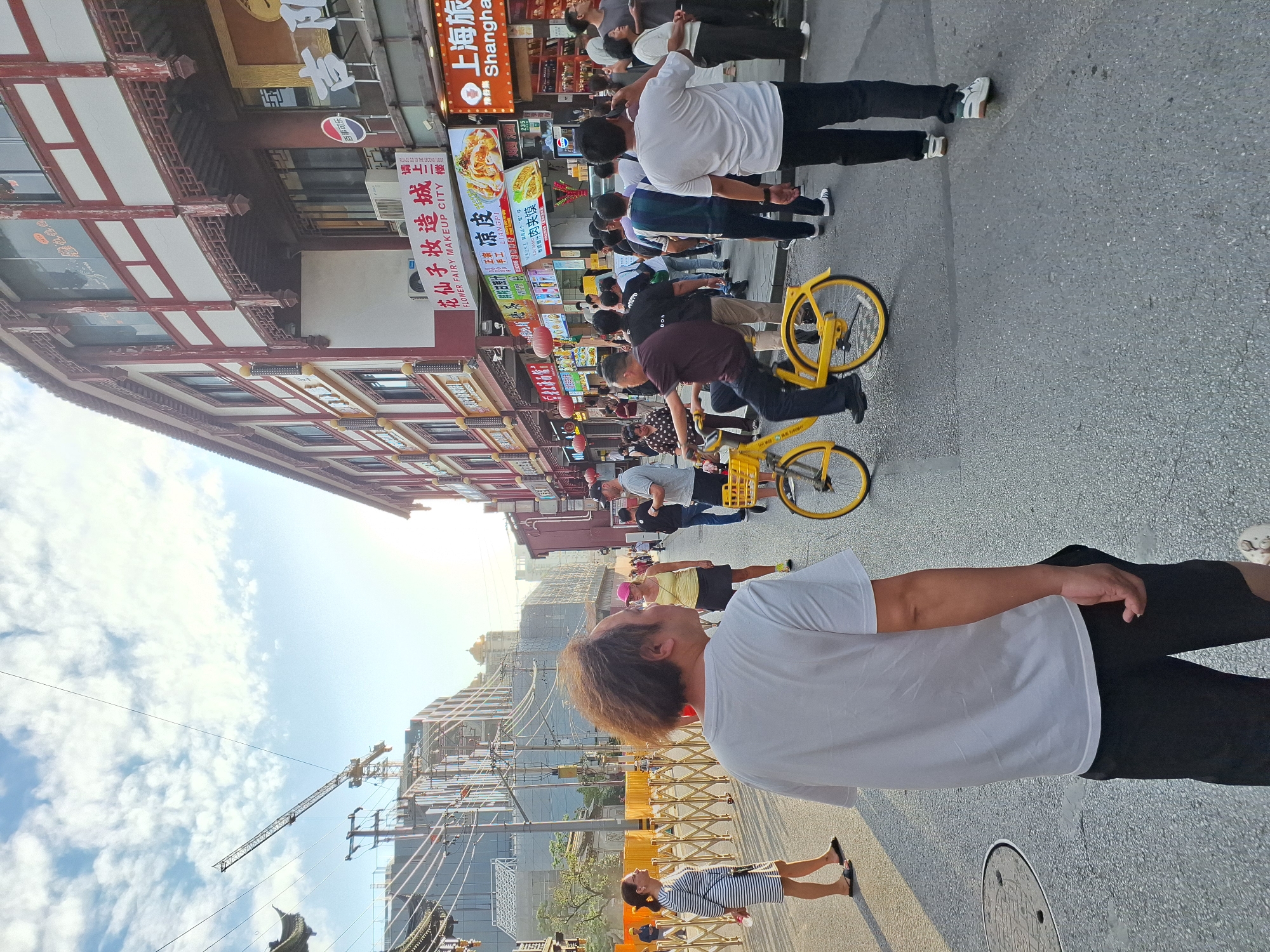 In Shanghai, China, a street scene unfolds with a mix of traditional and modern elements under a partly cloudy sky. The foreground features a man with disheveled hair wearing a white t-shirt and black shorts, facing away from the camera. To his left, a woman in a striped dress stands with her back to the camera. Further down the street, a man is riding a bright yellow bicycle with a basket. Several other pedestrians are visible, some walking, some standing, and some interacting with each other.
The background reveals a street lined with buildings exhibiting traditional Chinese architectural features, such as red pillars and layered roofs, interspersed with modern signage and storefronts. One prominent sign reads "FLOWER FAIRY MAKEUP CITY" in English. Construction cranes and scaffolding are visible in the distance, indicating ongoing development. The street itself is paved with asphalt, and streetlights and utility wires crisscross overhead. The overall atmosphere suggests a bustling urban environment in Shanghai.