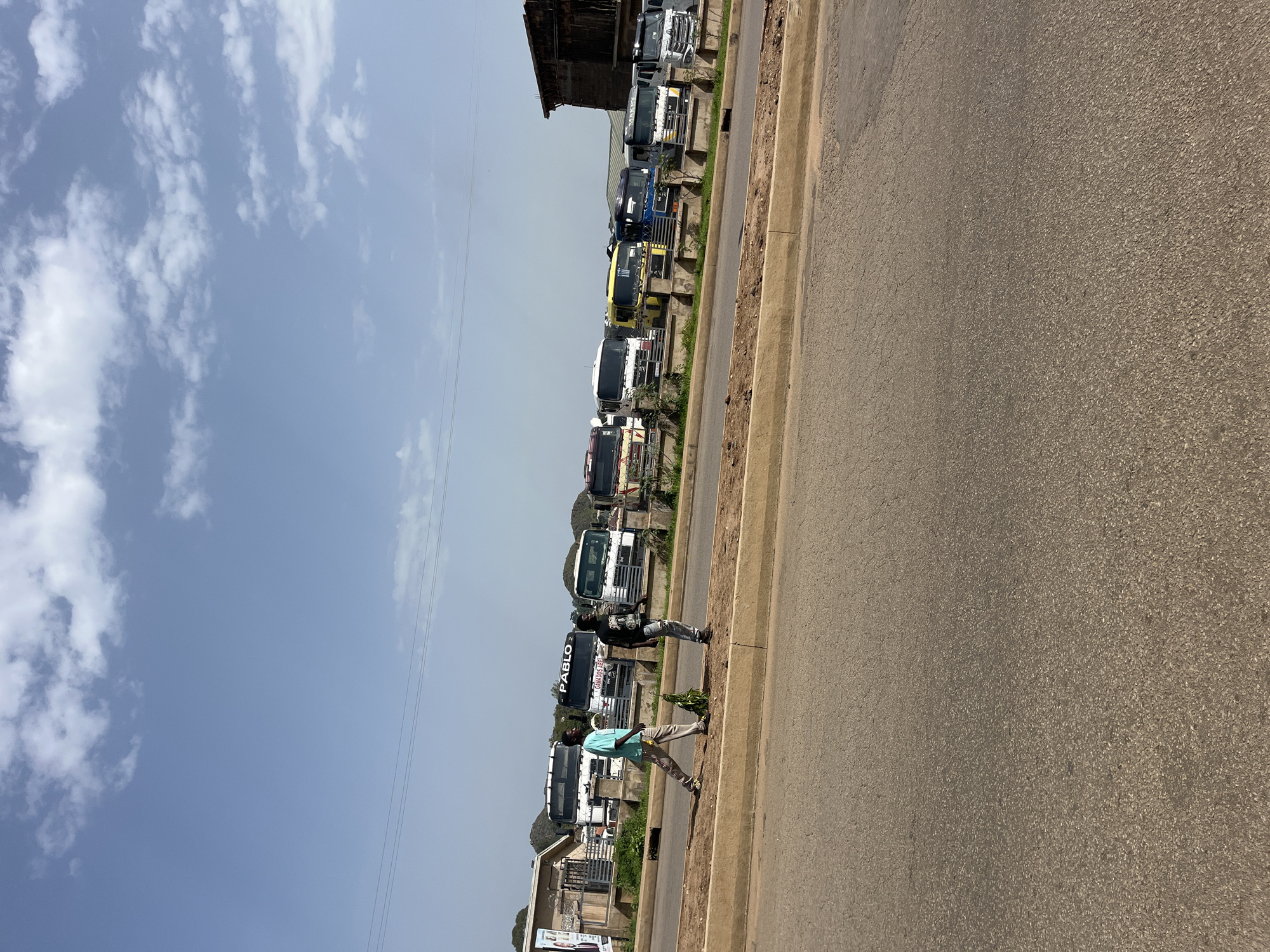Two men walk along a paved median next to a road in Bukuru, Nigeria. Behind them, a line of parked semi-trucks is visible. The sky is blue with scattered white clouds. The men appear to be walking in the same direction. The truck on the far left has the word "PABLO" written on it. One man is wearing a blue t-shirt and khaki pants, and the other is wearing a black t-shirt with graphics and ripped blue jeans. The environment is sunny.