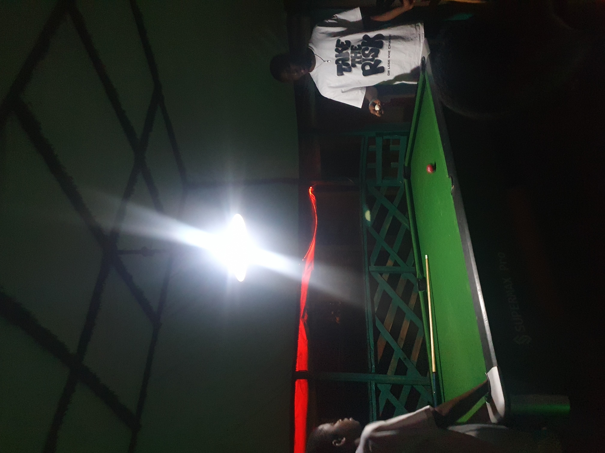 Two young Black males are playing billiards at night under a covered outdoor area. The room is dimly lit by a bright overhead light and a string of red lights along the far railing. The billiards table is green with a red ball and a cue stick visible. One male, wearing a white t-shirt with the words "TAKE THE RISK" printed on it, is standing on the right side of the table holding a white cue ball. The other male, wearing a light-colored t-shirt, is standing on the left side of the table. The location is Lokoja, Nigeria.