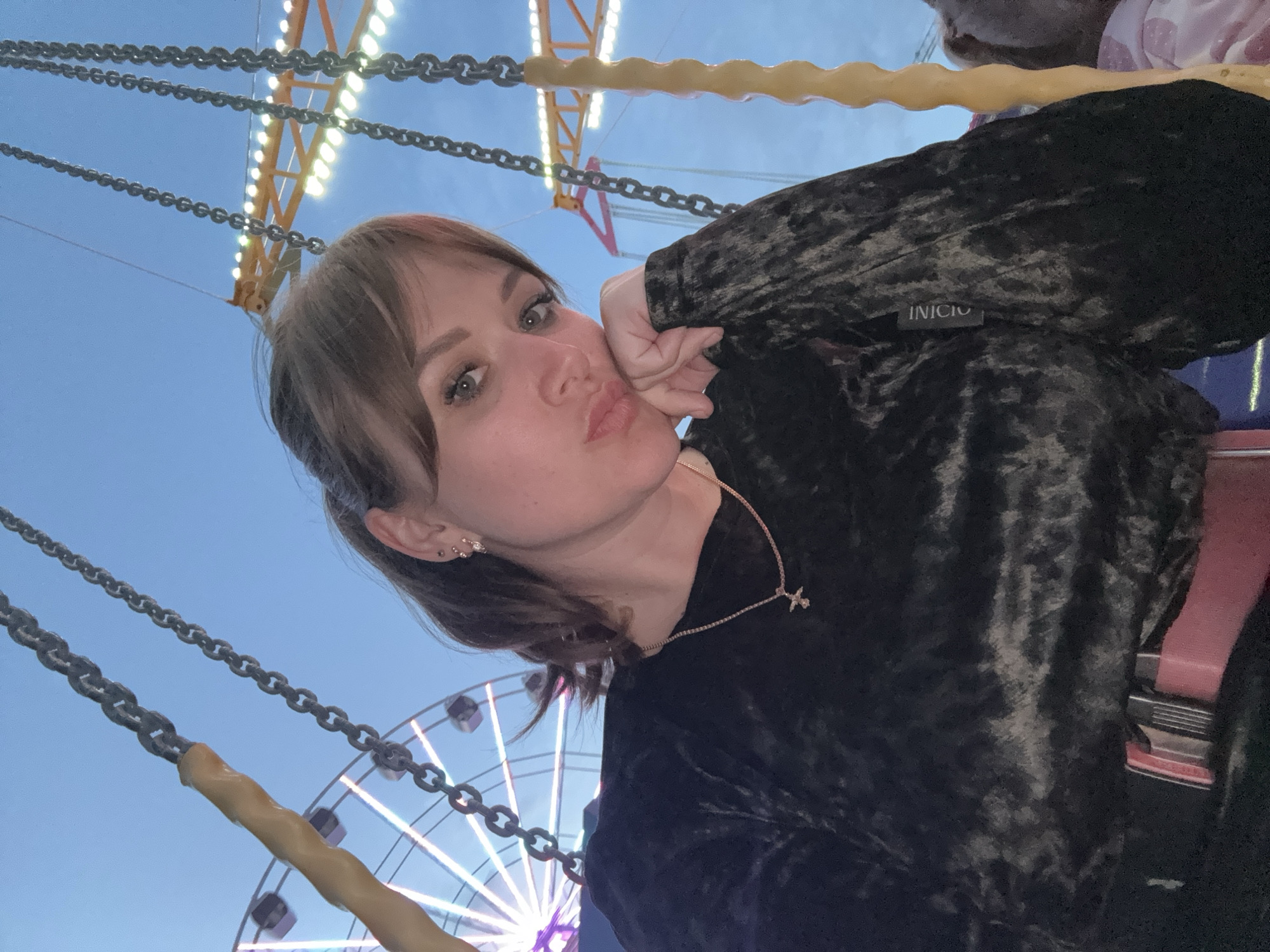 A young woman is photographed at an amusement park at dusk. The background features a swing ride with illuminated towers and a Ferris wheel, both typical amusement park attractions. The woman, positioned in the foreground, is wearing a dark-colored velvet top, a necklace with a pendant, and earrings. She is seated in a swing ride, secured with a red seat belt. Her left hand is gently touching her face, and she is posing with puckered lips for the camera. The image is taken at an angle, providing a view of the rides and the sky. The lighting suggests it's taken in the evening, with artificial lights illuminating the park rides against the darkening sky. The woman's clothing has a tag visible that says, "INICIO." The Ferris wheel in the background is illuminated with white lights.