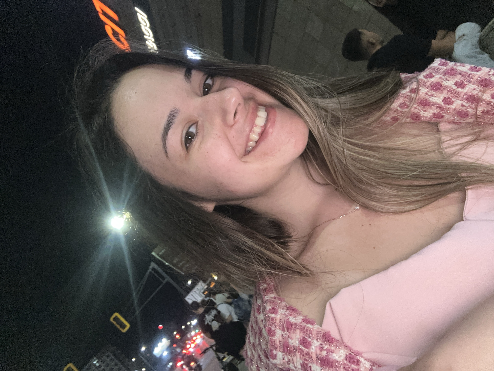 A young adult female, with light brown hair and brown eyes, is depicted in a close-up selfie taken at night in an urban environment in Sofia, Bulgaria. She is smiling broadly, revealing her teeth, and wearing a light pink top under a textured pink and white patterned jacket. A delicate silver necklace with a small pendant is visible on her chest. The background on the left features blurred red vehicle tail lights, white headlights, and indistinct figures, indicating street traffic and pedestrians. On the upper right, a partially visible, illuminated orange sign displays the letters "o" and "en", consistent with a "Technopolis" electronics store sign. A bright artificial light source in the upper left background creates a lens flare. The scene suggests a busy commercial district or a street adjacent to a retail establishment at night.