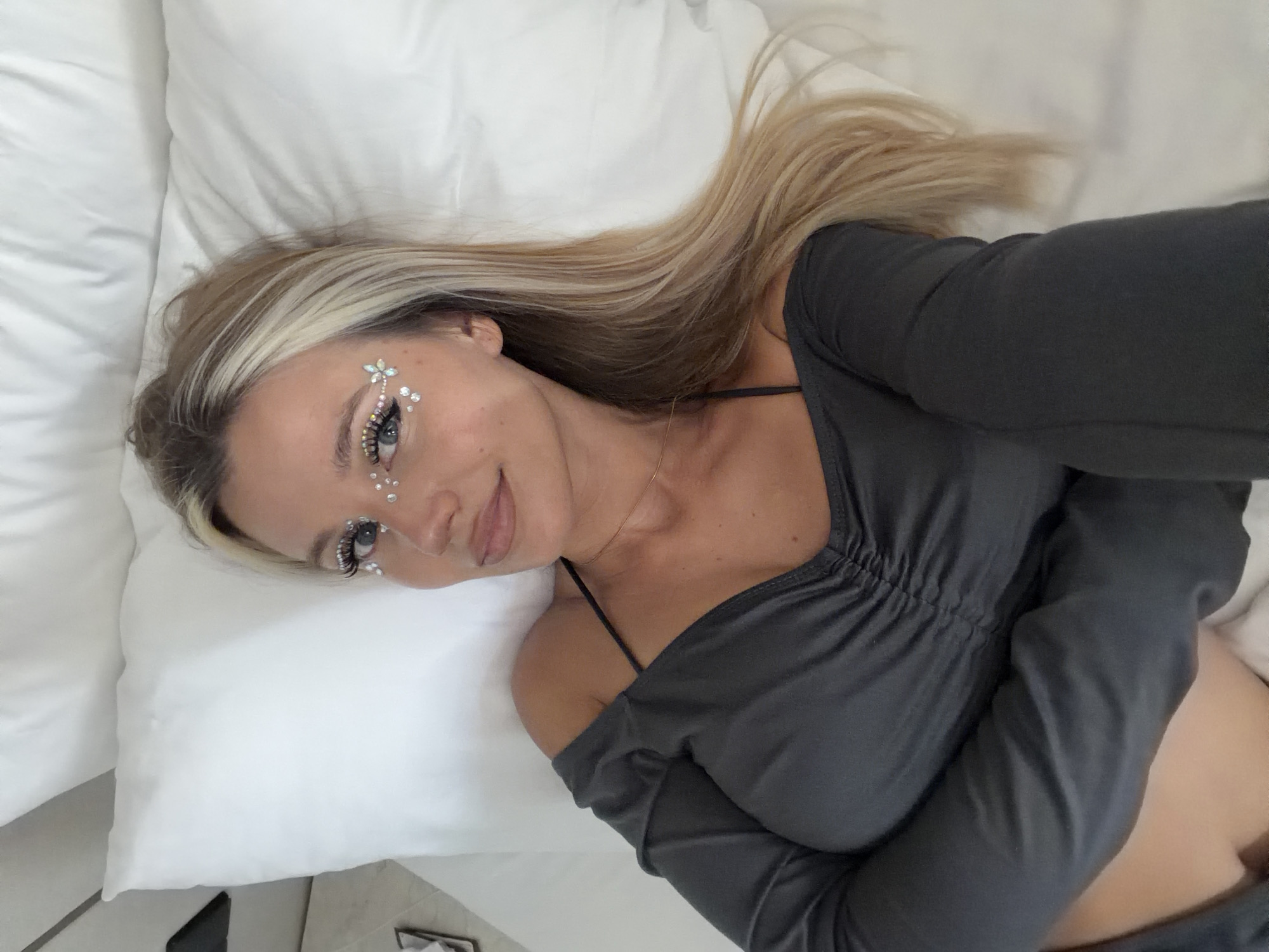 An overhead shot captures a young woman lying on a white pillow. She is adorned with sparkling face gems, some clustered around her eyes and others scattered across her cheekbones and forehead. Her blonde hair, streaked with lighter blonde highlights, is partially visible, flowing to the right of the frame. She wears a dark gray off-the-shoulder top with a ruched detail at the bust and a thin black strap visible. The background is dominated by a white pillow, suggesting an indoor setting, possibly a bed. The image appears to be a selfie taken from above. There are no visible landmarks or contextual clues to pinpoint the location in North Kuta, Indonesia.