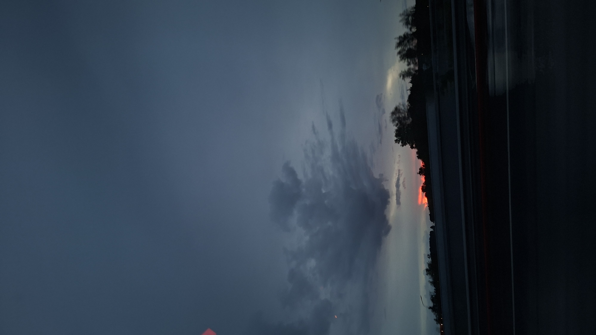 The image shows a view of a dark, cloudy sky at what appears to be dusk or dawn. There's a line of trees silhouetted against a faint red glow on the horizon, suggesting a sunrise or sunset. The right side of the image shows a dark, reflective surface, possibly a wet road.
The location is Лугаваслабадскі сельскі Савет, Belarus.