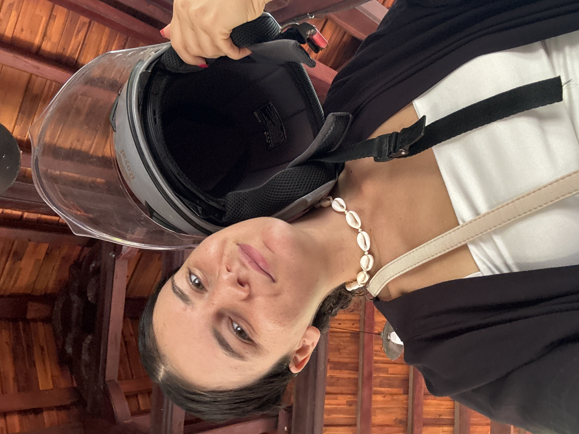 A young woman with dark hair and brown eyes, wearing a seashell necklace and a black cardigan over a white top, holds a motorcycle helmet by its chin strap. She is looking at the camera with a slight smile. The background shows a wooden ceiling with exposed beams, suggestive of an outdoor or semi-outdoor structure. The image appears to be taken in Mũi Né, Vietnam, possibly at a guesthouse or a roadside stall due to the rustic architecture.
