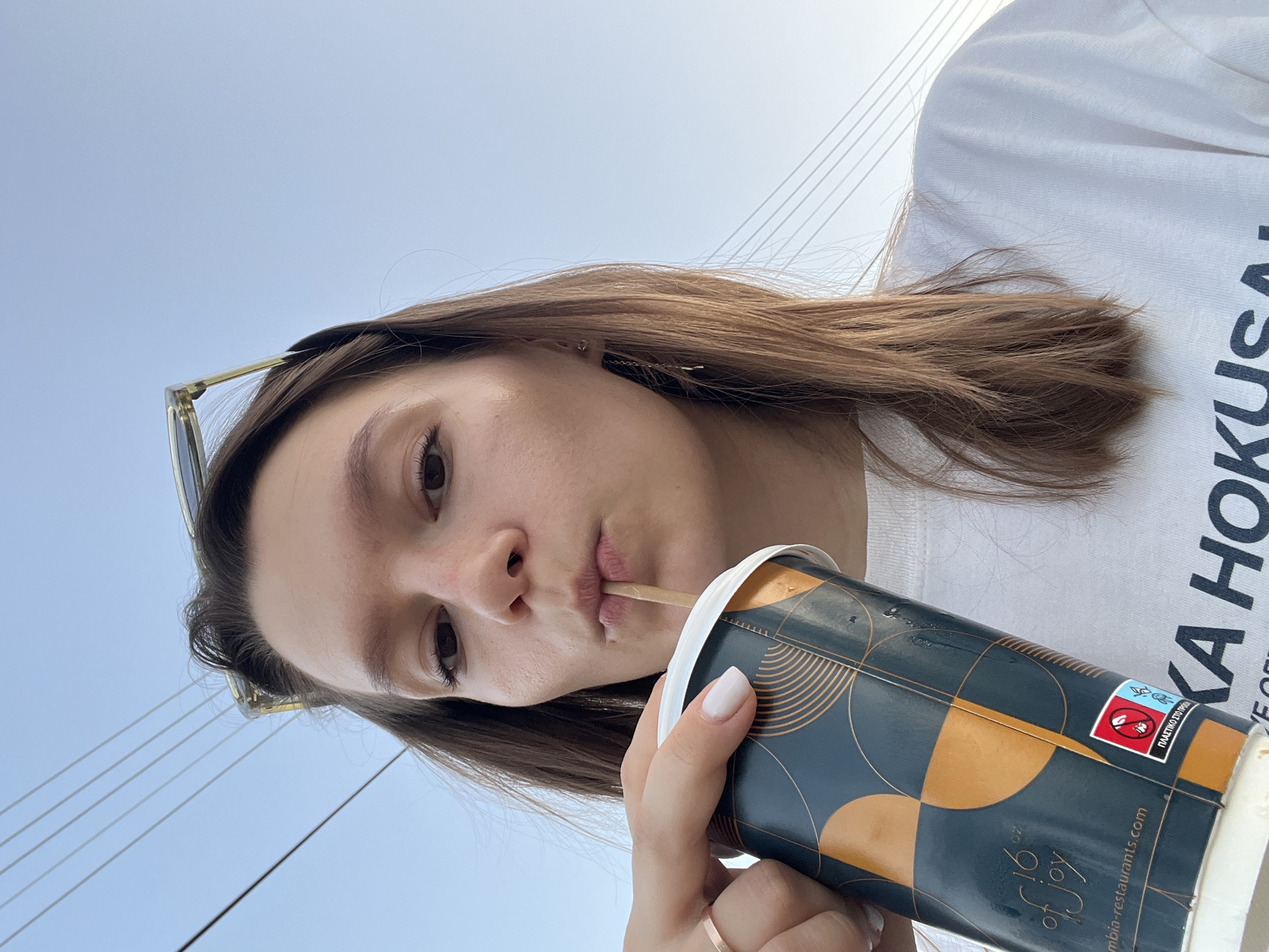 A young woman is taking a selfie outdoors, holding a large paper cup with a straw and drinking from it. She is wearing a white t-shirt and a ring on her left hand. Her sunglasses are pushed up on her head. In the background, the sky is visible with several power lines stretching across it. The cup has a patterned design and text, including "16 oz of joy" and "columbia-restaurants.com." There are also some Greek characters visible on a sticker on the cup. The lighting suggests it is daytime. Based on the Greek text and the presence of similar fast-food chains in the region, this image was likely taken in Limassol, Cyprus, possibly near a food establishment or a public outdoor area with overhead power lines.
