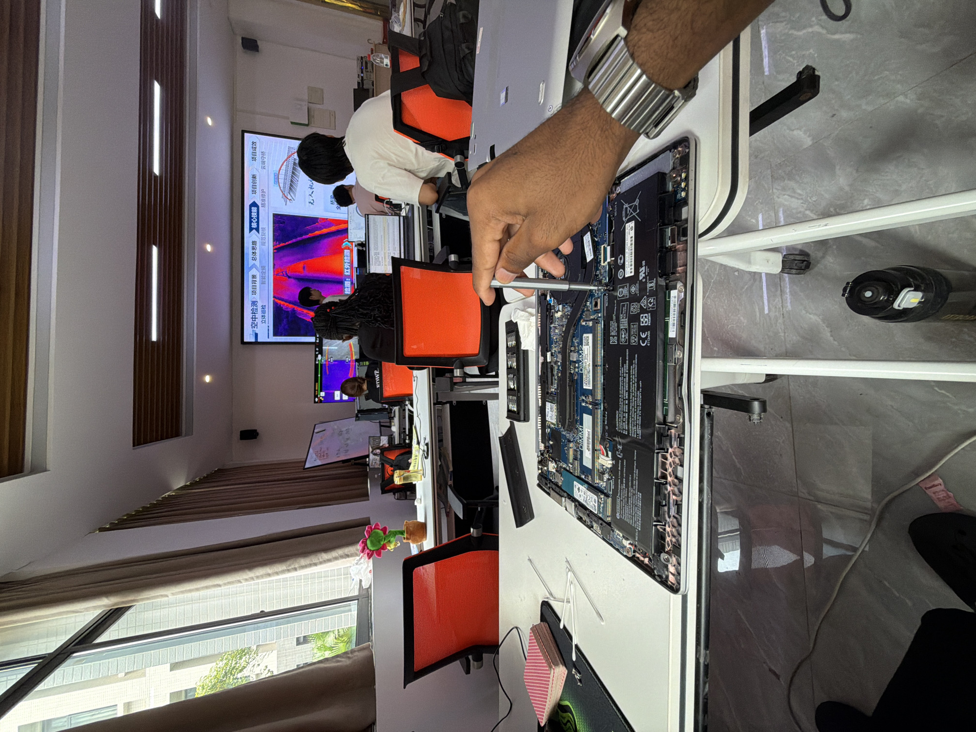 A first-person perspective shows a hand in the foreground using a silver screwdriver to work on the internal components of a disassembled laptop. The laptop's motherboard and battery are visible on a white desk. To the left of the laptop are a stack of playing cards on a black mousepad and some white plastic tools. In the background, several people are seated at desks in an office environment. A large screen displays an image of a road from a thermal camera and Chinese text. The room has large windows with beige curtains, and red office chairs are visible. A potted plant with a pink flower sits on a desk near a window. The scene appears to be an office in Jiangjin, China, likely a technology or engineering workspace.