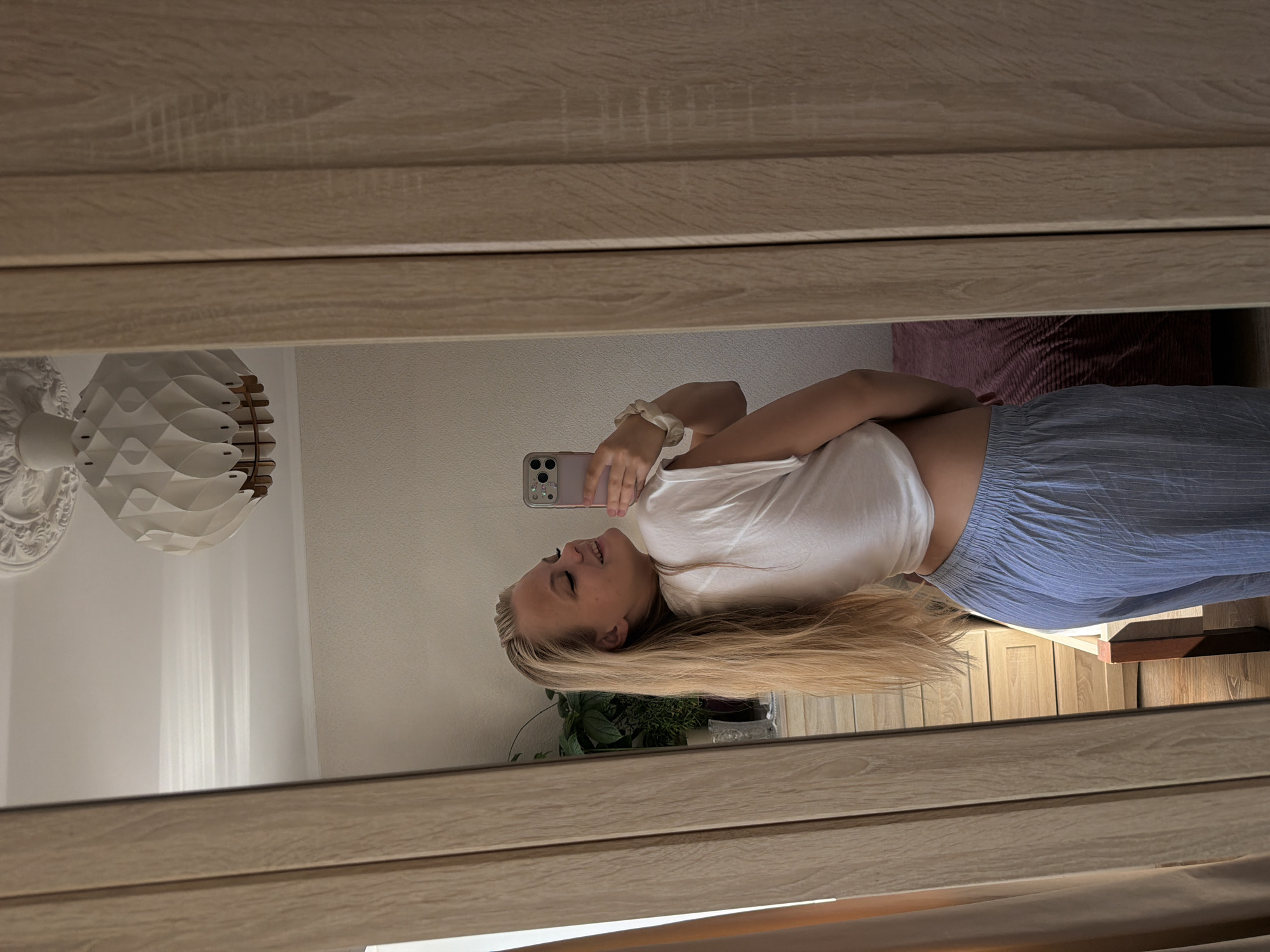 A young woman with long blonde hair is taking a selfie in a mirror. She is wearing a white t-shirt and blue striped pajama bottoms. She is standing in what appears to be a bedroom, with a wardrobe to her right and a dresser with a plant on it in the background. The room is brightly lit by natural light. There is a modern, decorative ceiling light fixture visible at the top of the frame. The image is taken from a slightly low angle, and the woman is looking over her shoulder at the camera, smiling. The woman's body is not fully visible as she is turned away from the camera and the image cuts off at her knees. The presence of a wardrobe and dresser suggests this is a domestic setting. The image does not contain any elements that would allow for identification of the specific location within the city of Chaikovsky, Russia.