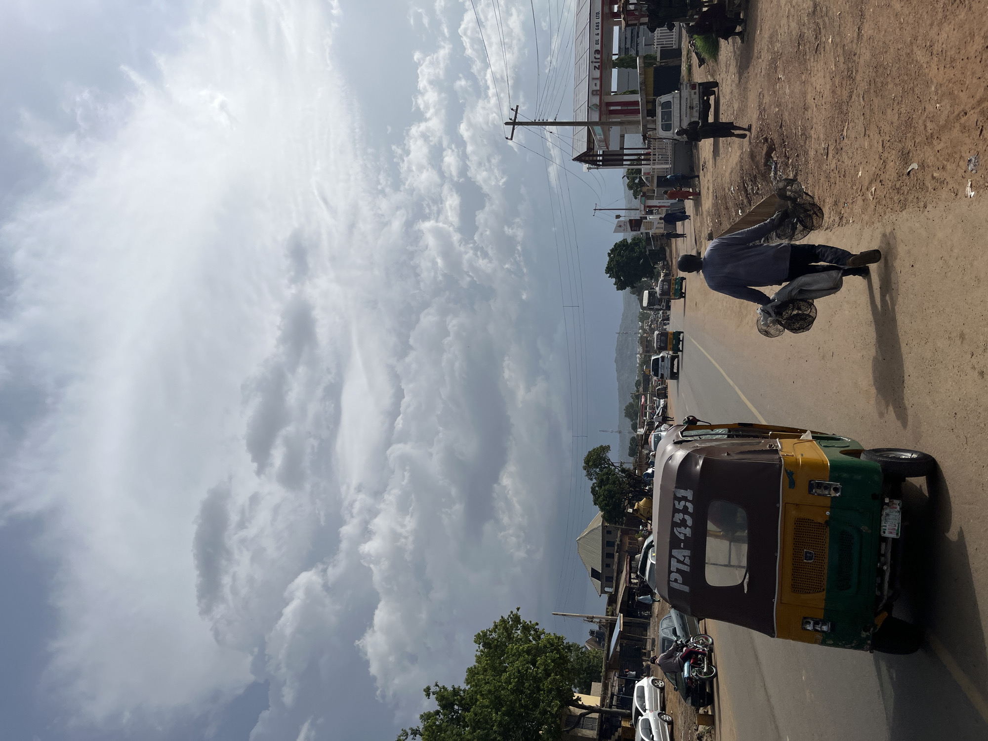 The image depicts a street scene in Jos, Nigeria, under a heavily clouded sky. In the foreground, a man walks away from the camera carrying several large, circular wire mesh items. To his left, a yellow and green auto-rickshaw with the license plate PTA-4351 is parked on the side of the road. Further down the street, several other vehicles are visible, including cars, a truck, and another auto-rickshaw. Buildings line the right side of the road, including a gas station with a prominent sign that reads "Zira Oil & Gas Ltd." and another building with what appears to be solar panels on its roof. A few other individuals are visible walking along the roadside or near the buildings. The ground is a mix of asphalt road and dirt. The overall environment suggests a busy, unpaved urban area.