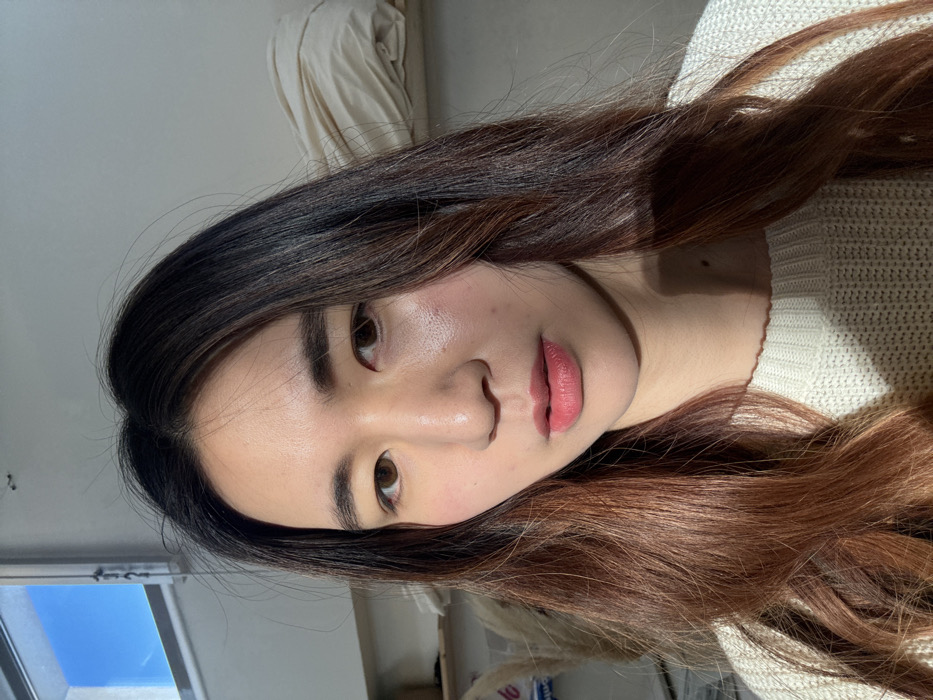 A medium close-up portrait depicts an East Asian female indoors in Arcueil, France. The subject, appearing to be in her twenties, faces forward with a direct gaze towards the camera. She has long, dark brown hair transitioning to reddish-brown ombré ends, brown eyes, and fair skin with evident highlights on the forehead, nose, and cheeks from a strong light source positioned to the upper right. Her lips are colored with pink lipstick. She wears an off-white, textured knit sweater. The background consists of a plain, light-colored wall. A white-framed window showing clear blue sky is visible in the upper left quadrant. To the upper right, behind the subject's right shoulder, a stack of neatly folded, light-colored fabric rests on a shelf or elevated surface. Additional obscured objects, including what appears to be dried botanical decor, are partially visible on the left side of the frame.