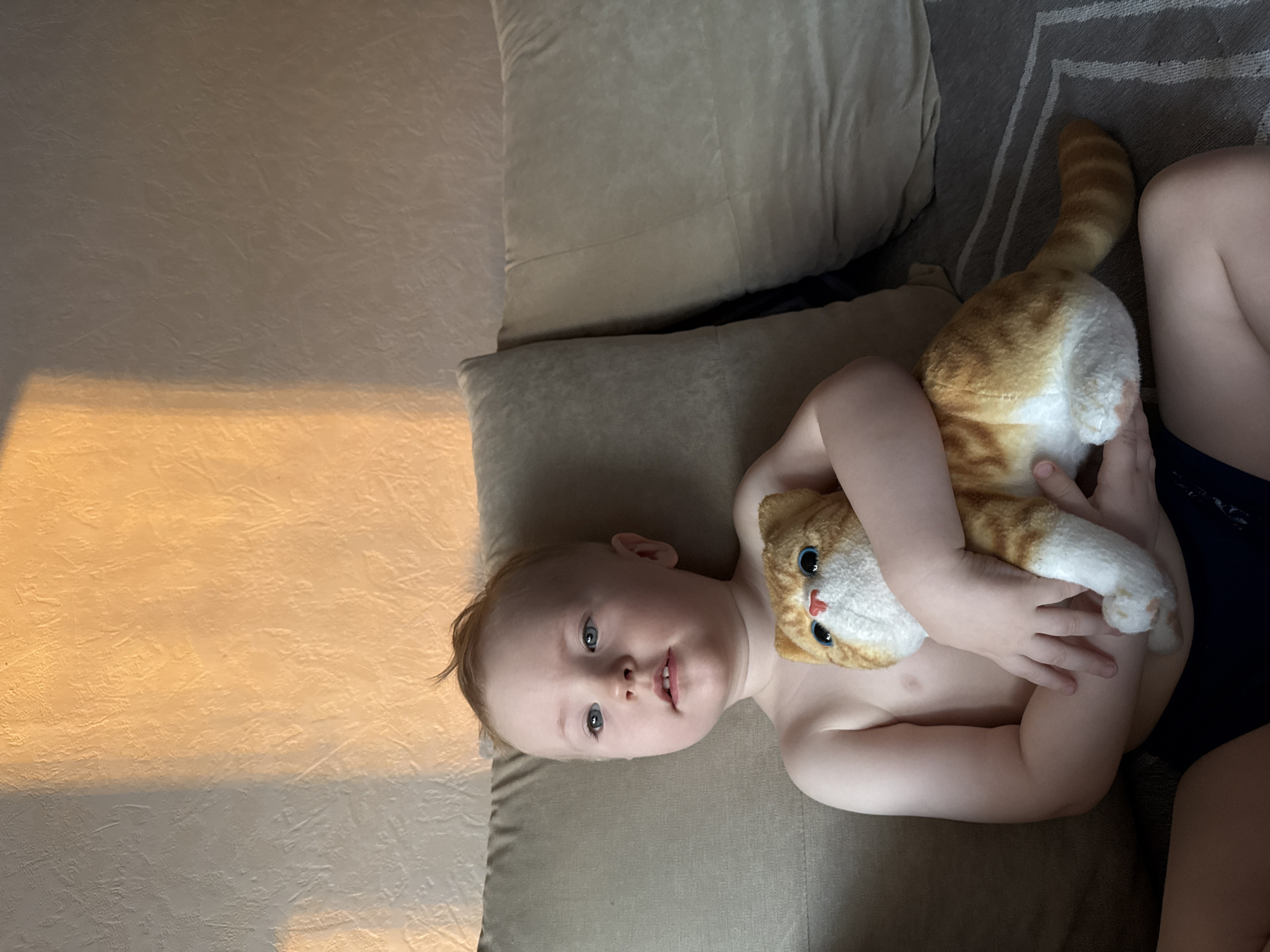 A child lies on a light brown cushion with a stuffed, orange and white tabby cat. The child has fair skin, blue eyes, and blonde hair. Part of a beige wall with a light shining is to the left, and the edge of a gray carpet is visible in the top right. The scene is indoors. There is no information to suggest the location of this picture is Chaikovsky, Russia.