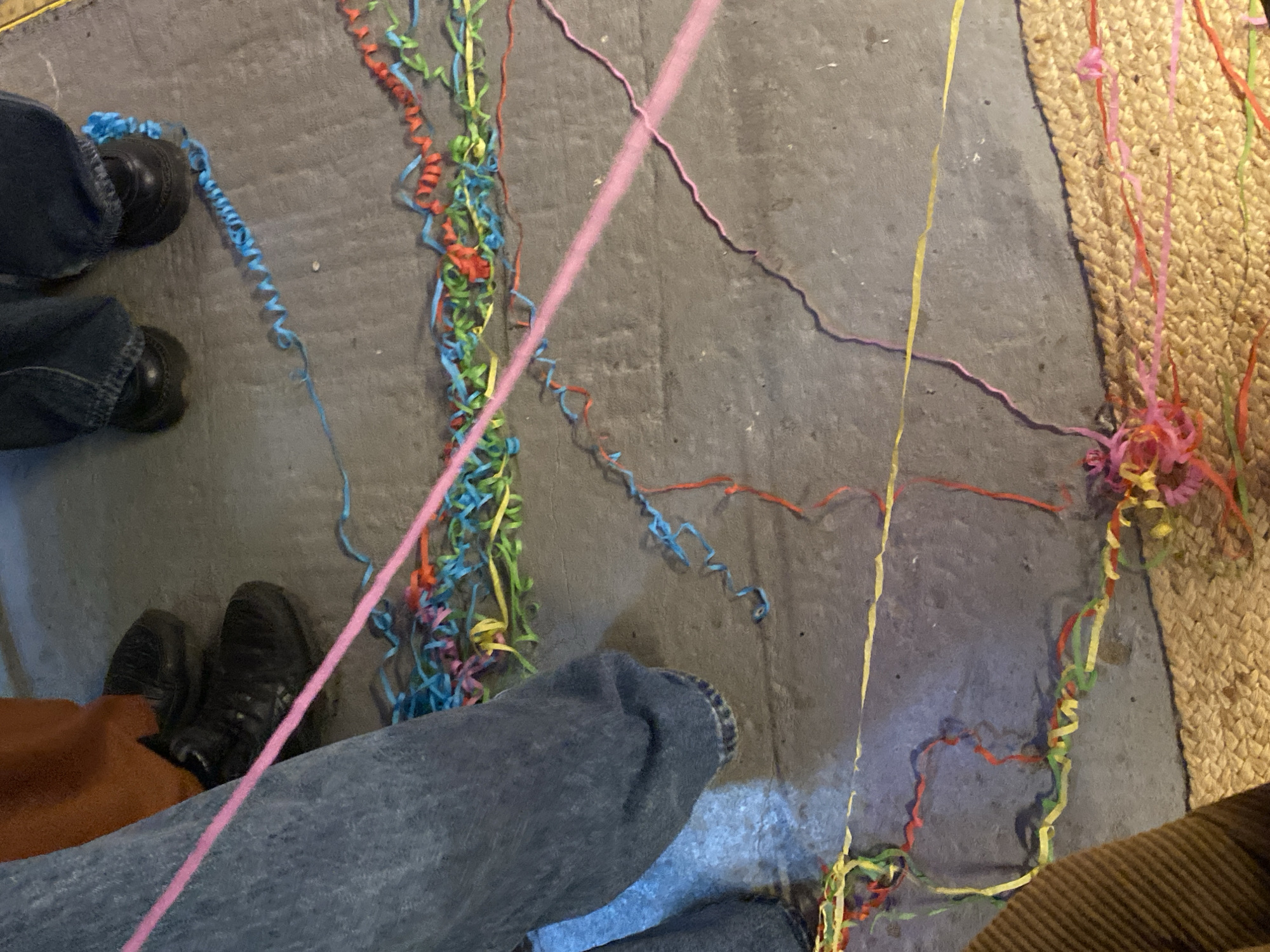 The image depicts a scene on a gray concrete floor covered with colorful party streamers and confetti. Two pairs of feet wearing black shoes are visible on the left side of the image. A pair of legs wearing blue jeans is situated in the lower center. The upper right corner reveals a portion of a woven rug. No information is available regarding the location in the city of Заволжье, Russia.