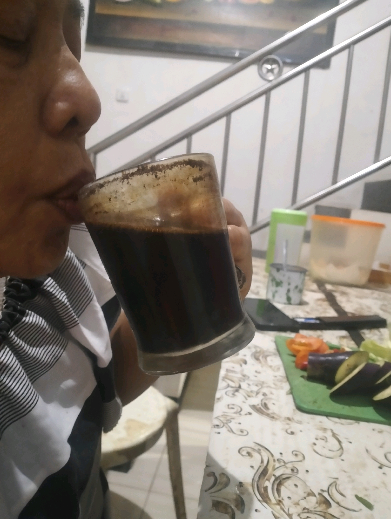 A close-up, medium shot depicts a person drinking from a glass of dark liquid. The person, seen from the nose down, is wearing a striped shirt. The glass has some residue on the rim. In the background, a stainless steel railing is visible, suggesting an indoor staircase. On a table in the foreground, a cutting board with sliced eggplant and tomatoes is present, along with other containers and a mobile phone. The environment appears to be a domestic setting. The presence of vegetables suggests food preparation might be underway. The location is identified as Klecorejo, Indonesia.