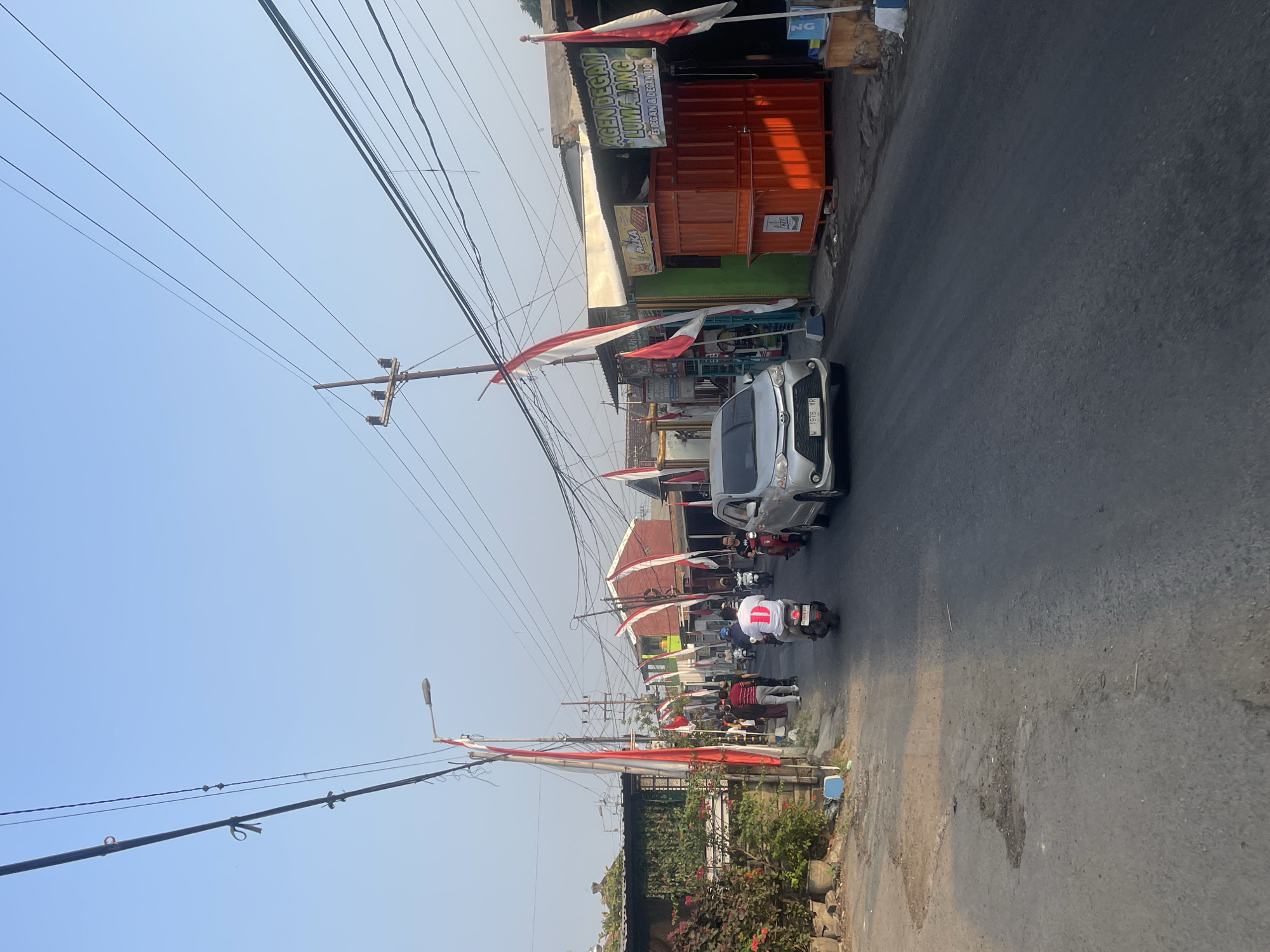 The image depicts a street scene in Kedungkendo, Indonesia, characterized by a road lined with buildings and adorned with numerous Indonesian flags. The flags, predominantly red and white, are mounted on poles along the roadside and strung across power lines, indicating a festive or patriotic occasion.

In the foreground, the asphalt road shows signs of wear, with some patched areas. On the left side of the road, there is a sidewalk area with potted plants and a stone wall. On the right side, a row of small shops and buildings is visible, one of which is an orange kiosk with signage indicating it is an "AGEN DEGAN LUMAJANG," selling coconut drinks.

Several vehicles are present, including a silver sedan with the license plate "W 1976 VR" in the foreground and several motorcycles. People are seen riding the motorcycles and walking on the side of the road. The sky is clear and blue, suggesting it is daytime, likely late afternoon given the angle of the sunlight casting shadows. Utility poles with a complex network of wires are prominent overhead.