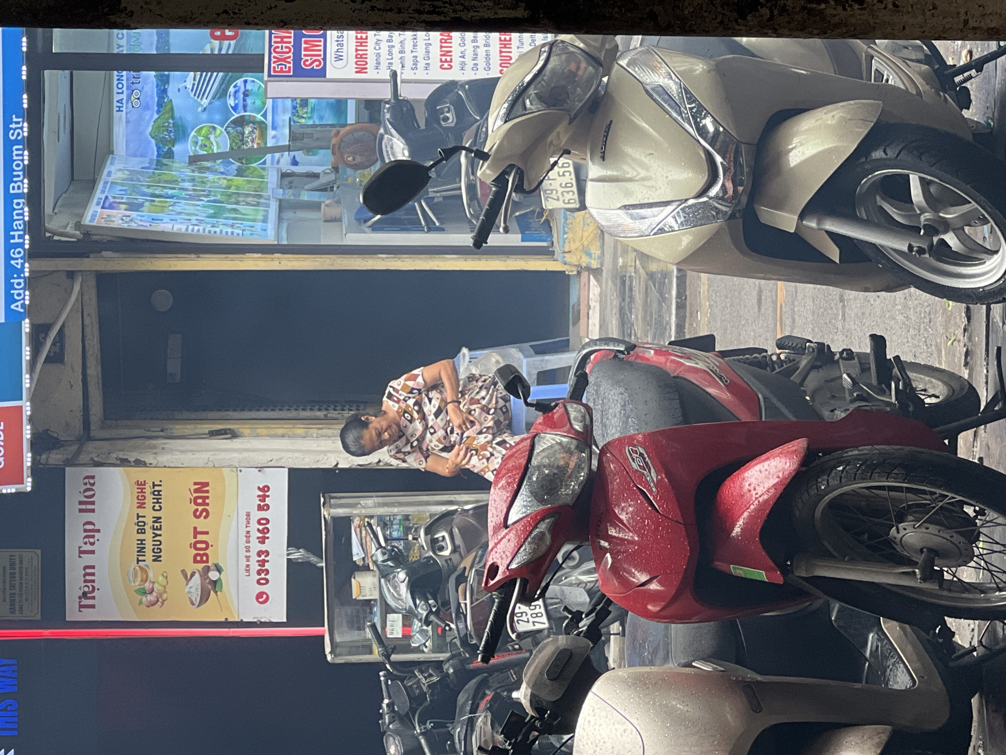 The image shows a street scene in Hanoi, Vietnam, likely in the Old Quarter based on the architecture and density of businesses. In the foreground, several parked motorcycles, some wet from rain, are visible. A woman in a patterned dress sits on a blue stool in front of a shop, appearing to be busy with something in her hands. Behind her, a shop window displays travel brochures for locations like Ha Long Bay. The shop appears to be a travel agency and possibly a small convenience store, as suggested by the signage which includes "Tiệm Tạp Hóa" (grocery store) and travel-related advertisements. The overall atmosphere is that of a busy urban street with a mix of commerce and daily life.