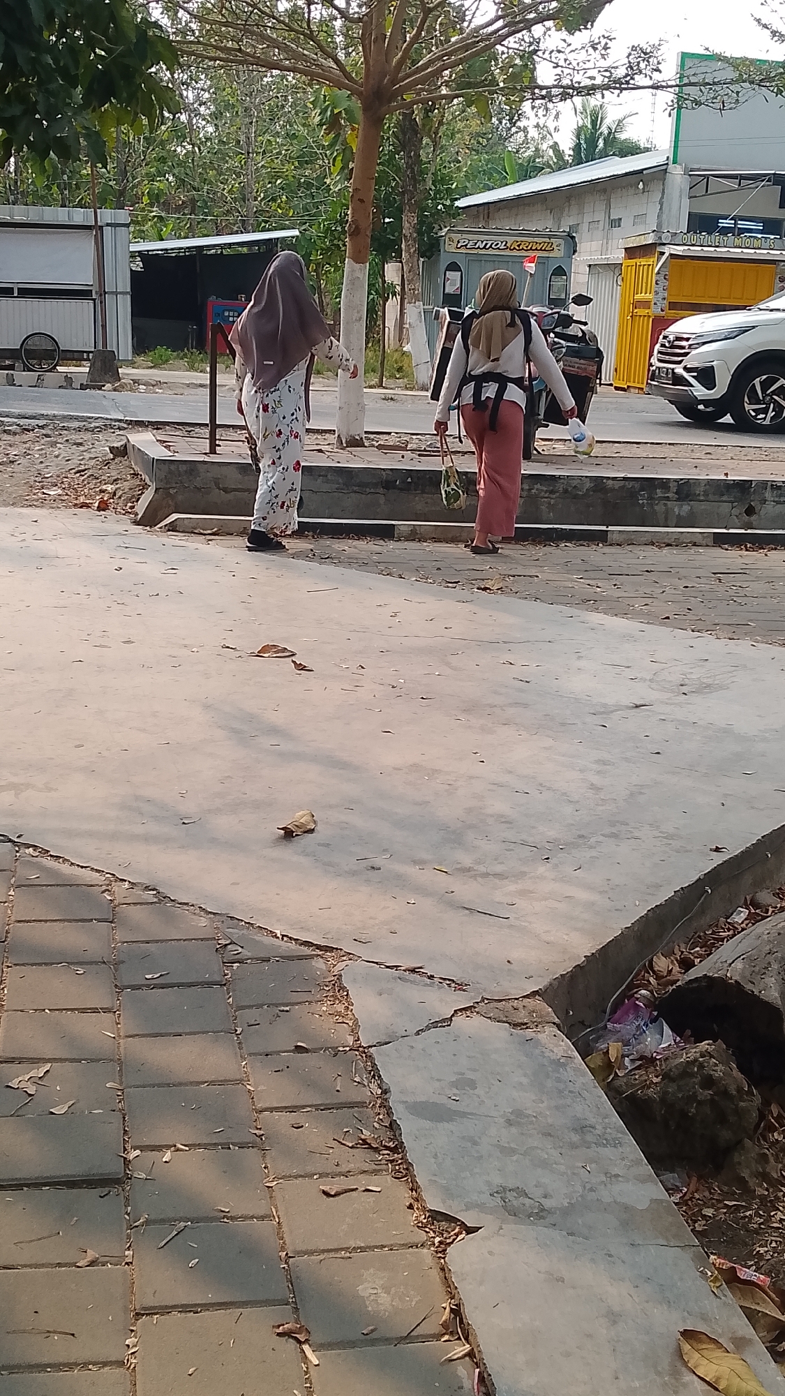 Two women wearing hijabs are walking on a sidewalk in what appears to be a commercial area. The woman on the left is wearing a floral-patterned dress and a dark brown hijab. The woman on the right is wearing a peach-colored hijab, a white shirt, and pink pants. She is carrying a bag over her shoulder and has a baby carrier on her back. In the background, there are several buildings, including what looks like a food stall with a sign that says "PENTOL KRIMIL" and a white car parked on the street. The sidewalk is made of concrete slabs and pavers, with some debris and leaves scattered around. The area seems to be in Karanglangu, Indonesia, given the presence of local Indonesian businesses.