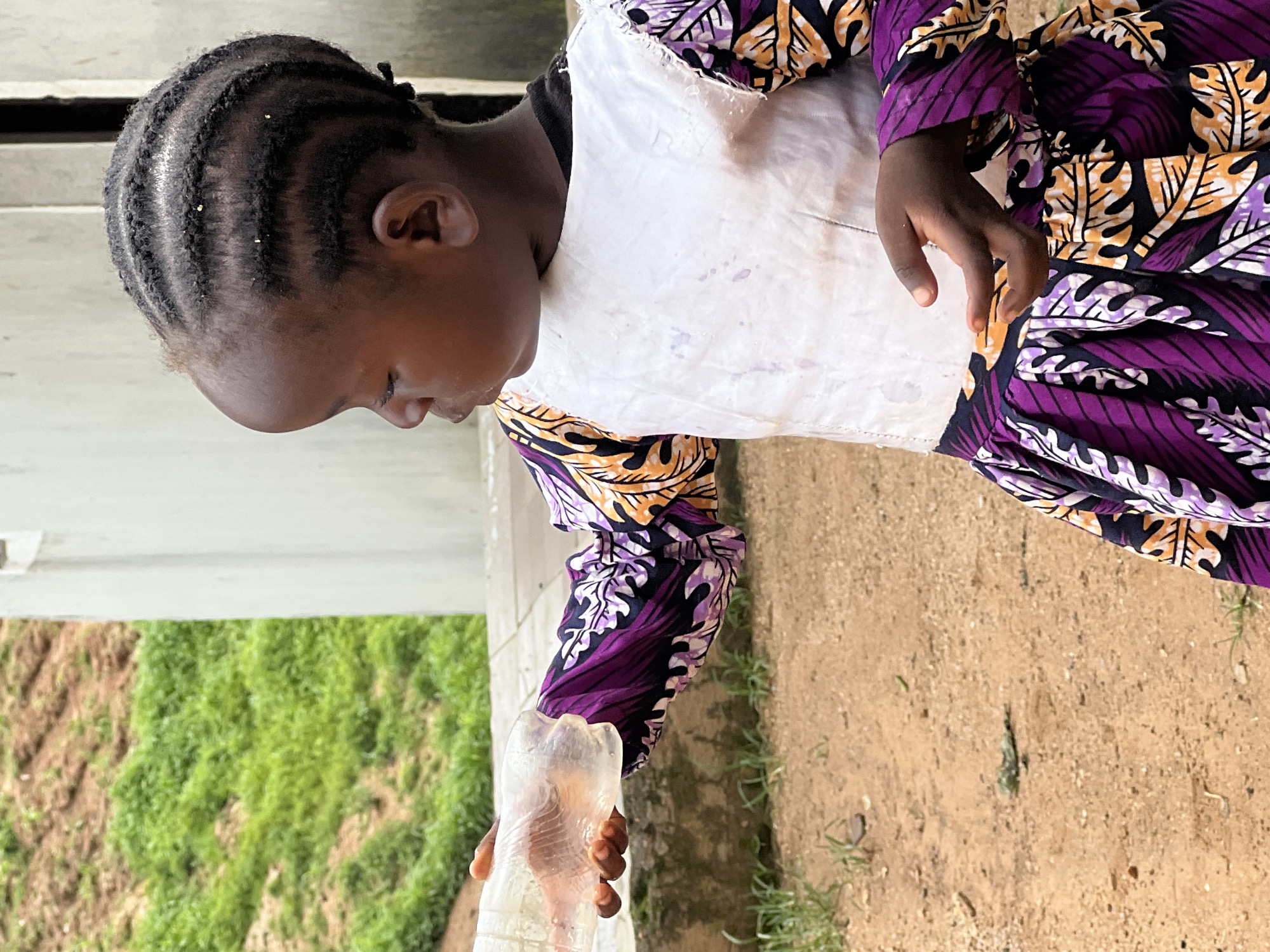 The image shows a young Black girl with braided hair, wearing a white overlay over a vibrant purple, orange, and black patterned dress. She is standing outdoors, with a dirt and gravel ground beneath her. In the background, there is a white building and a grassy slope with some terraced earth. The girl is holding a clear plastic water bottle, offering it out of frame to her right. Her left hand is extended slightly towards the bottle. The lighting suggests it is daytime. This scene is likely located in Jos, Nigeria, based on the context provided.