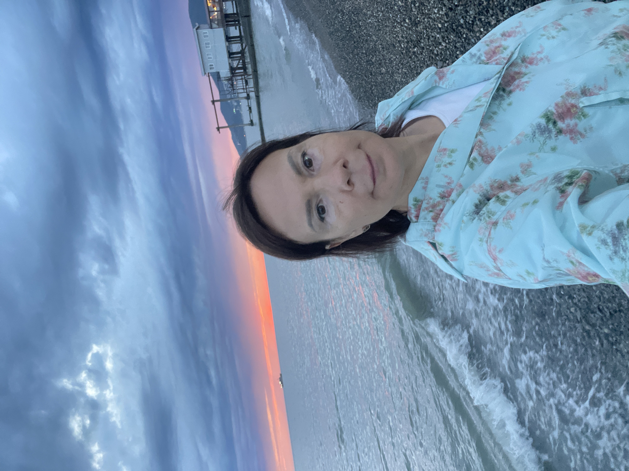 A medium shot depicts a woman, appearing to be in her 40s, wearing a floral light-blue jacket and a white t-shirt. She is positioned on a pebble beach with waves reaching the shore. Behind her, a structure resembling a pier or platform extends over the water. The background features a sky with a prominent sunset gradient, transitioning from deep blue to orange hues. The scene is potentially located in Sochi, Russia, given the description.