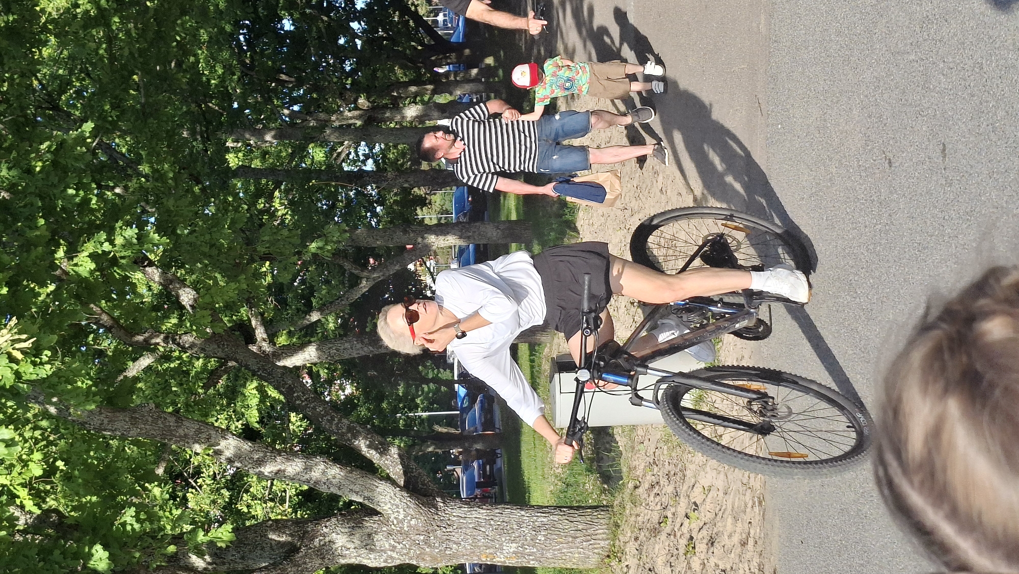 A woman with blonde hair is riding a bicycle on a paved path next to a grassy area with trees. She is wearing a white long-sleeved shirt, black shorts, and white sneakers. She has sunglasses on and is touching her ear. In the background, a man in a striped shirt and denim shorts is walking with a young child wearing a red hat and a patterned shirt. The man is holding a paper bag. They are walking on the same path as the woman on the bicycle. Several cars are parked in the background. The scene appears to be a park or a scenic area in Tallinn, Estonia, given the architecture and vegetation which are characteristic of the region.