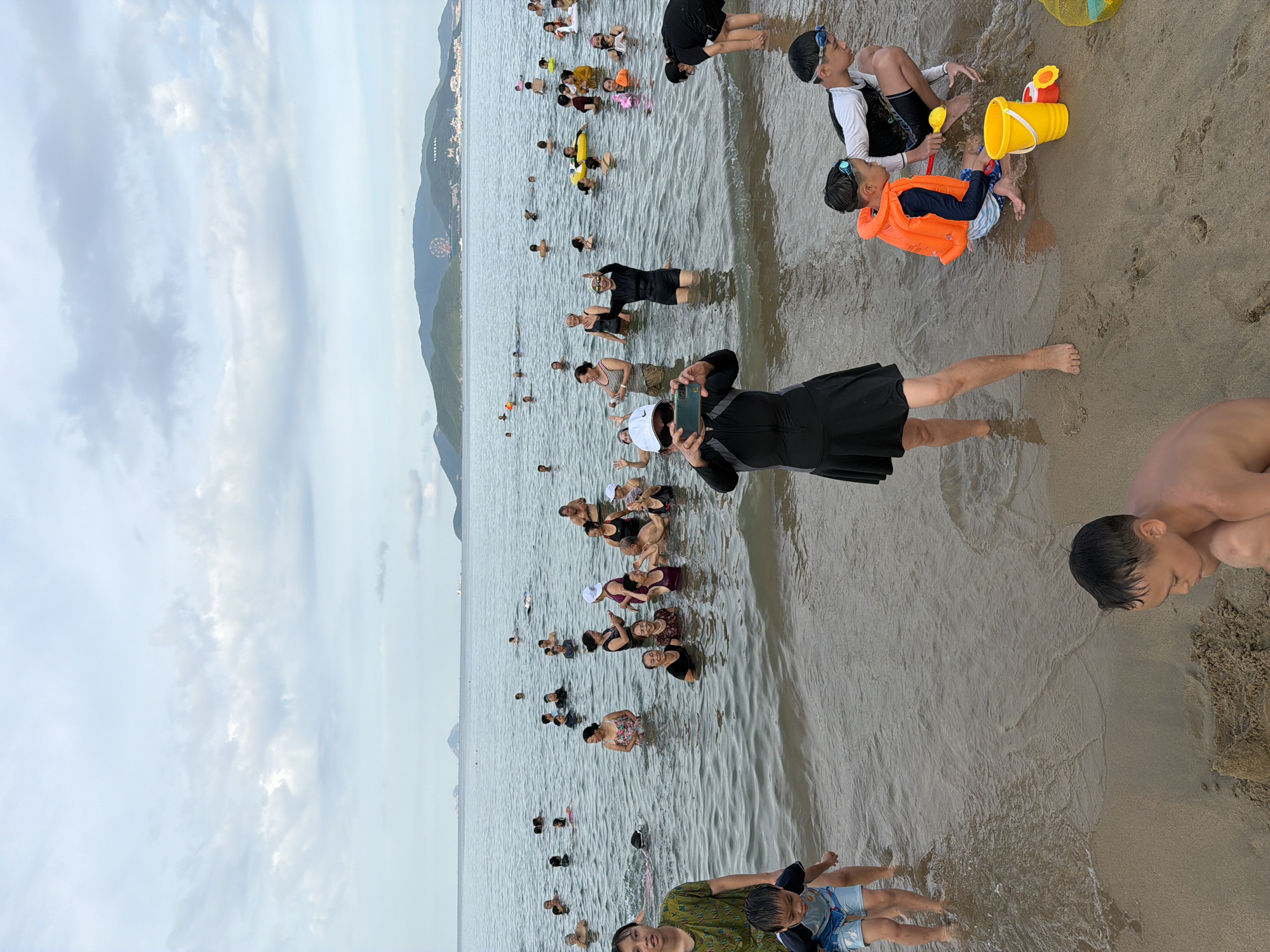 This is a medium shot of a crowded beach in Nha Trang Ward, Vietnam, on a cloudy day. Many people, including adults and children, are in the shallow water, some swimming and others standing or sitting. The water is calm and the beach is sandy. In the background, there are mountains and a Ferris wheel.
In the foreground, on the sand, two young boys are playing with sand toys. One boy is wearing an orange life vest and the other is wearing swim trunks. To the left of the boys, a woman in a black swimsuit is standing and holding a phone, taking a picture of the scene. Further back in the water, there are many groups of people interacting, with some swimming and others talking. The overall atmosphere is one of leisure and recreation.