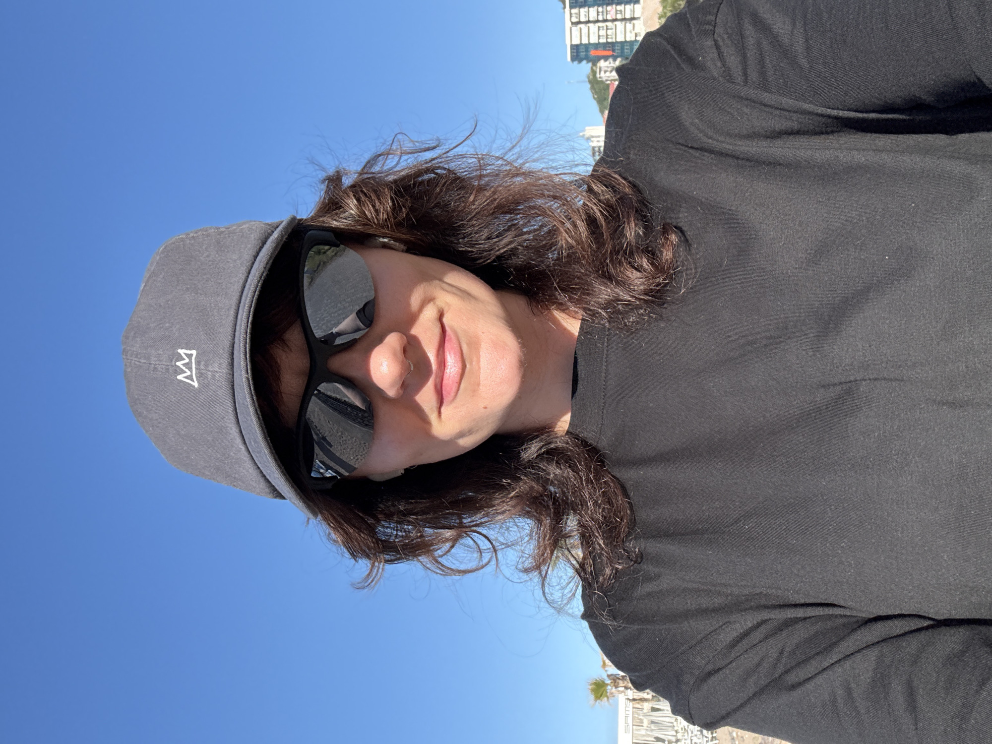 The image shows a person wearing a dark grey baseball cap, black sunglasses, and a black long-sleeved shirt. The person is smiling slightly. The sunglasses reflect a beach scene. The background features a clear blue sky and buildings, possibly apartments or hotels, located along the coast in Boreti, Montenegro.