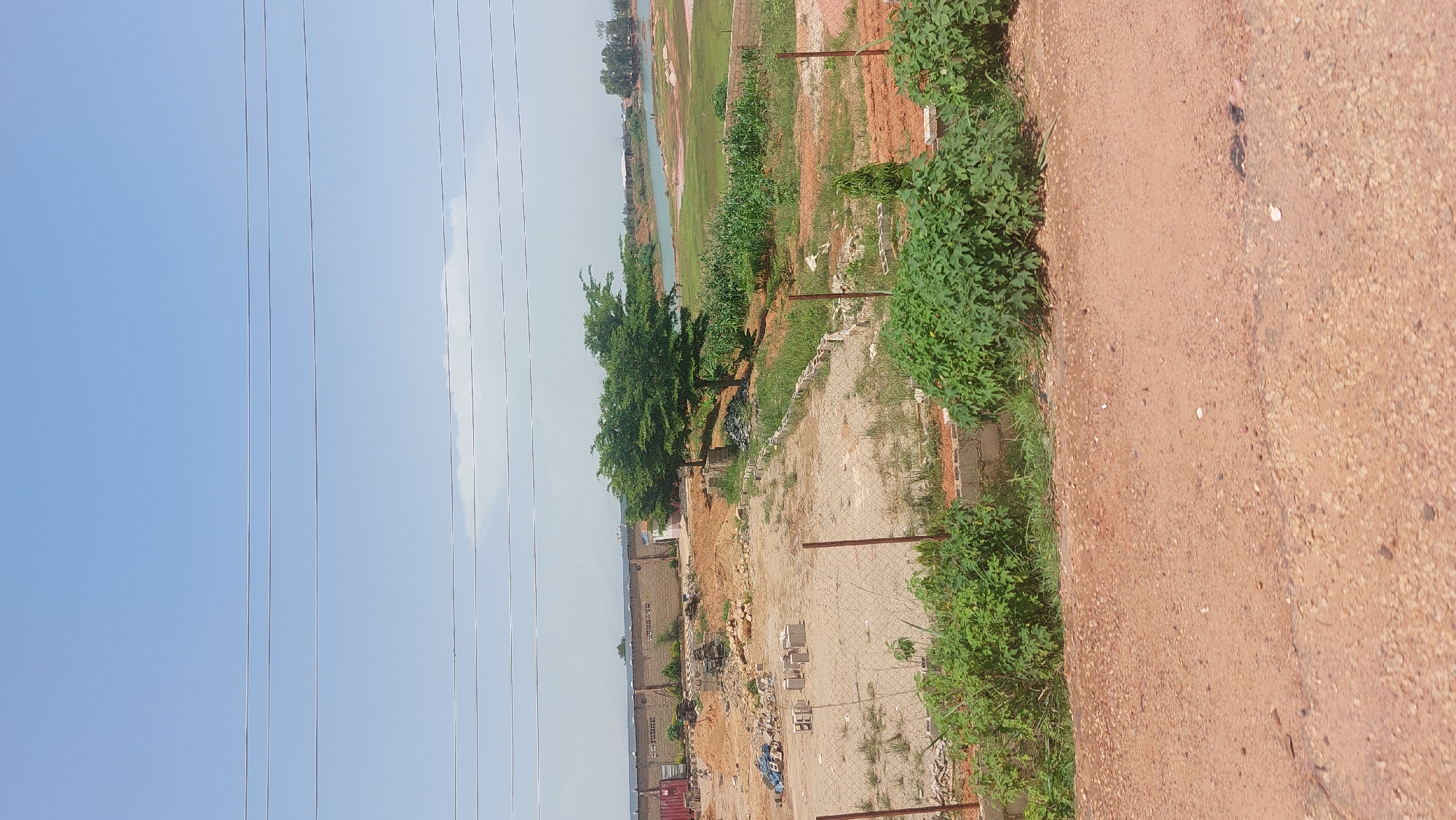 The image depicts an outdoor scene in Bar Jirgi Summa, Nigeria, featuring a construction site in the foreground and a body of water in the background. The foreground is dominated by a dirt road and a partially developed construction area with piles of bricks and building materials. A chain-link fence runs across the middle of the scene, separating the dirt road from the construction area. In the mid-ground, a single tree with a wide canopy stands near a brick wall that borders a body of water. Beyond the water, a natural landscape with vegetation and distant structures is visible. The sky above is clear and blue with faint white clouds. The scene appears to be under natural daylight.