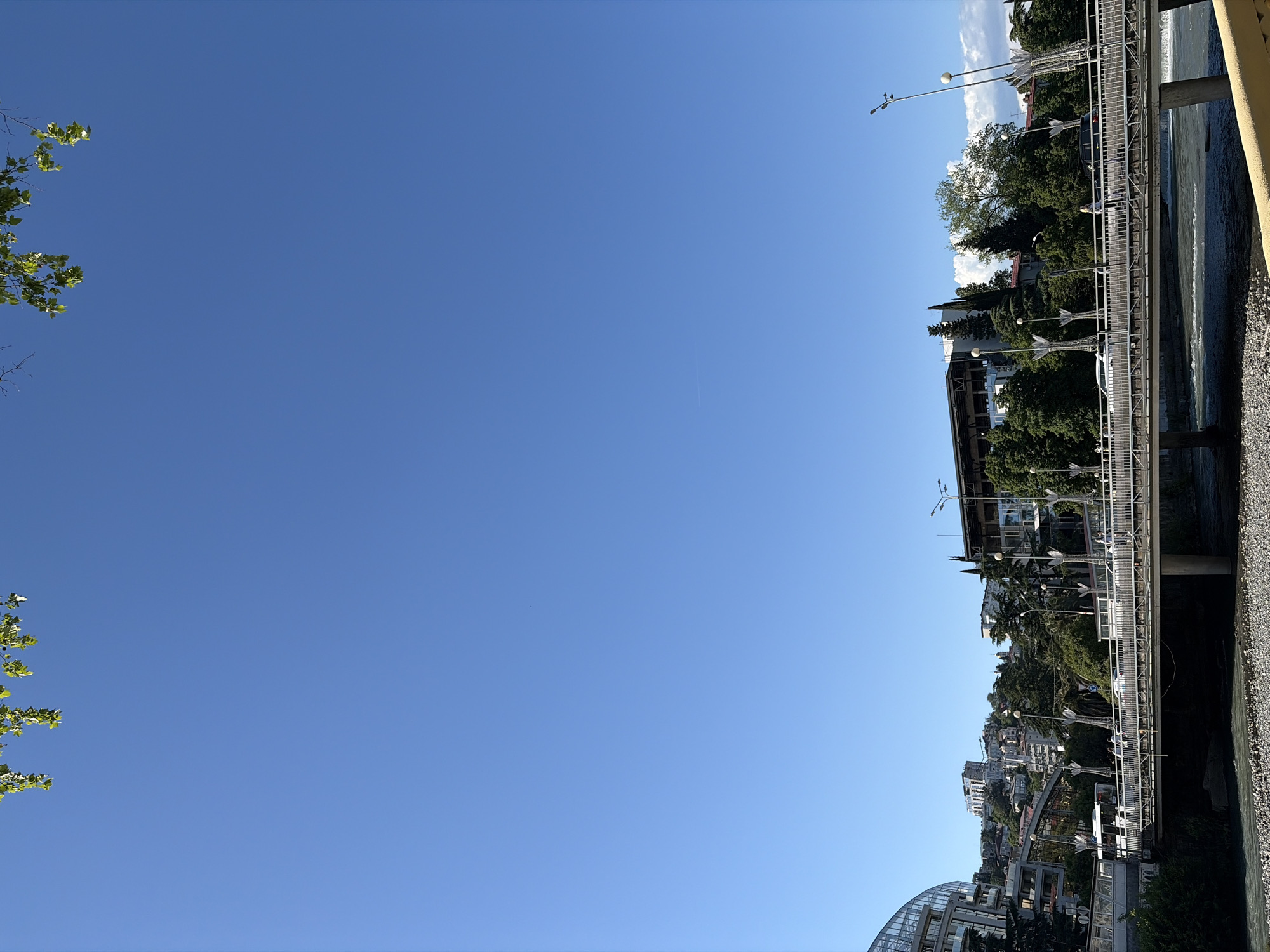 The image displays an outdoor urban landscape under a vast, clear blue sky. On the left, green leaves and branches frame the scene, suggesting the presence of a tree. The right side is dominated by a river or canal, with dark, rippling water and a pebbly bank in the foreground.
Running parallel to the waterway is a substantial metal bridge or elevated walkway, equipped with railings and numerous distinct lampposts. These lampposts feature multi-directional light fixtures, some appearing arrow-shaped. A dark vehicle and a person in light-colored clothing are faintly visible on the bridge. Beyond this structure, a cityscape stretches into the distance, comprising various buildings of different architectural styles nestled among dense, lush green trees and foliage, indicative of a hilly terrain. A modern building with a prominent curved glass roof is partially visible in the lower right corner.
The scene is captured in bright daylight, likely midday or early afternoon, under clear and sunny weather conditions. A faint contrail is visible in the expansive blue sky. The overall impression is one of a calm, everyday urban environment, with elements suggesting the subtropical and hilly landscape of Sochi, Russia. No specific event is taking place, and no visible text is present in the image.