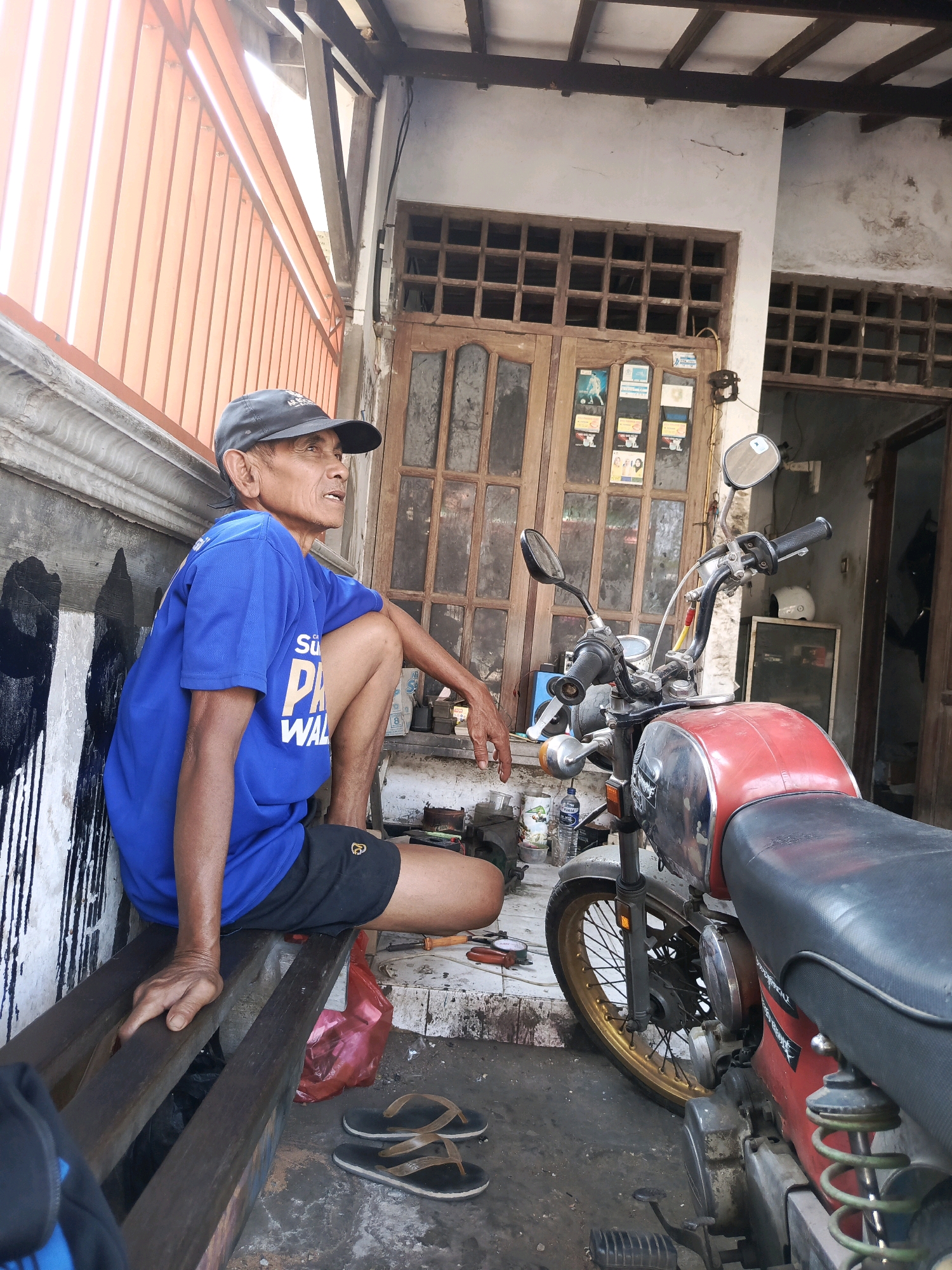 A man wearing a blue t-shirt and a black baseball cap is sitting on a wooden bench next to a red motorcycle. He is looking to his right, with his left leg bent and his right leg extended. The man appears to be in his 60s. The motorcycle is a vintage model with a chrome gas tank and black seat. The scene appears to be in an outdoor workshop, possibly in Semanding, Indonesia, with a wooden door and windows in the background. The ground is concrete, and there are tools and a red plastic bag on the ground. The overall impression is one of a rural or urban workshop setting.