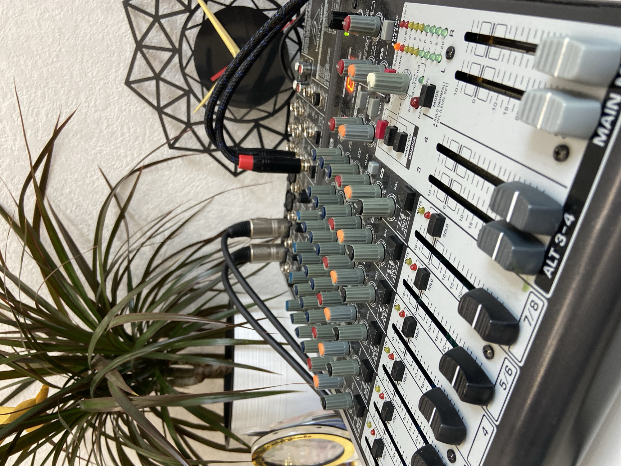 A close-up, eye-level, medium shot captures a portion of a white and gray Behringer Xenyx mixer with numerous colorful knobs and sliders. Cables are plugged into the top of the mixer. To the left, a potted Dracaena plant with long, green and red leaves stands in a white pot. A circular, metallic object with a yellow edge is partially visible behind the plant. To the right of the mixer, a black geometric wall clock with yellow and red hands is mounted on a white, textured wall. The scene appears to be indoors. There are no people present. The lighting is bright and natural.