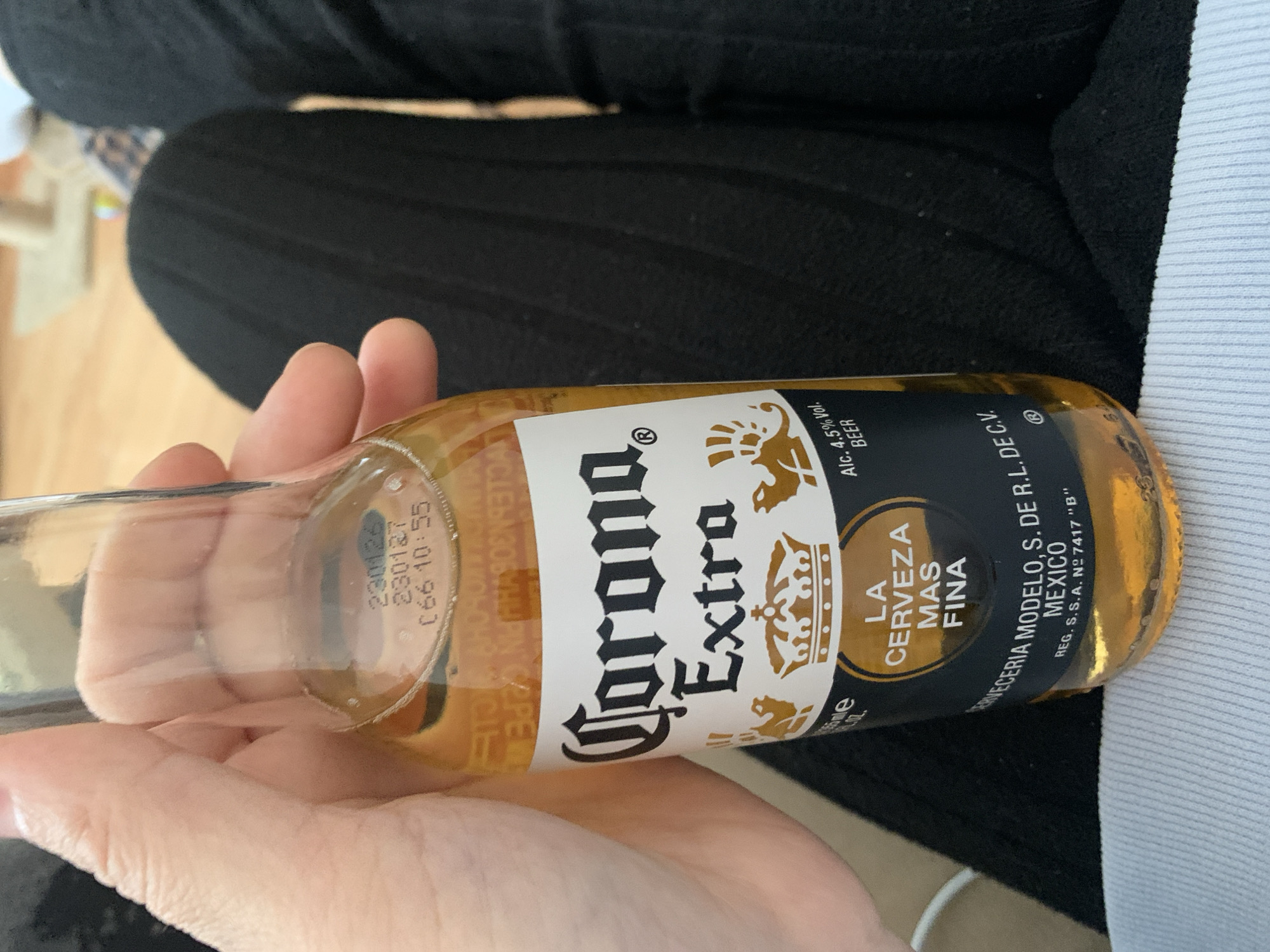 A person's hand holds a bottle of Corona Extra beer in an indoor setting in Stavropol, Russia. The image is taken at a slight angle, with the hand and bottle positioned against a background of dark, possibly black, fabric which appears to be clothing or upholstery. A lighter, blurry surface, potentially wood flooring, is visible in the upper left, and a section of light blue ribbed fabric is seen on the right side of the frame.
The clear glass bottle contains a light amber beer. The label is white with black text and gold accents, prominently displaying "Corona Extra" and the slogan "LA CERVEZA MAS FINA" (The Finest Beer). Other visible text on the label includes "Alc. 4.5% Vol.", "BEER", "CERVECERIA MODELO. S. DE R.L. DE C.V.", "MEXICO", and "REG. S.S.A. Nº 7417 "B"". Near the neck of the bottle, a laser-etched code is visible on the glass, showing "230126", "230127", and "C66 10:55", which likely pertains to production or expiration information. The activity shown is simply holding the bottle, with no specific event or discernible emotions conveyed. The lighting suggests an indoor environment.