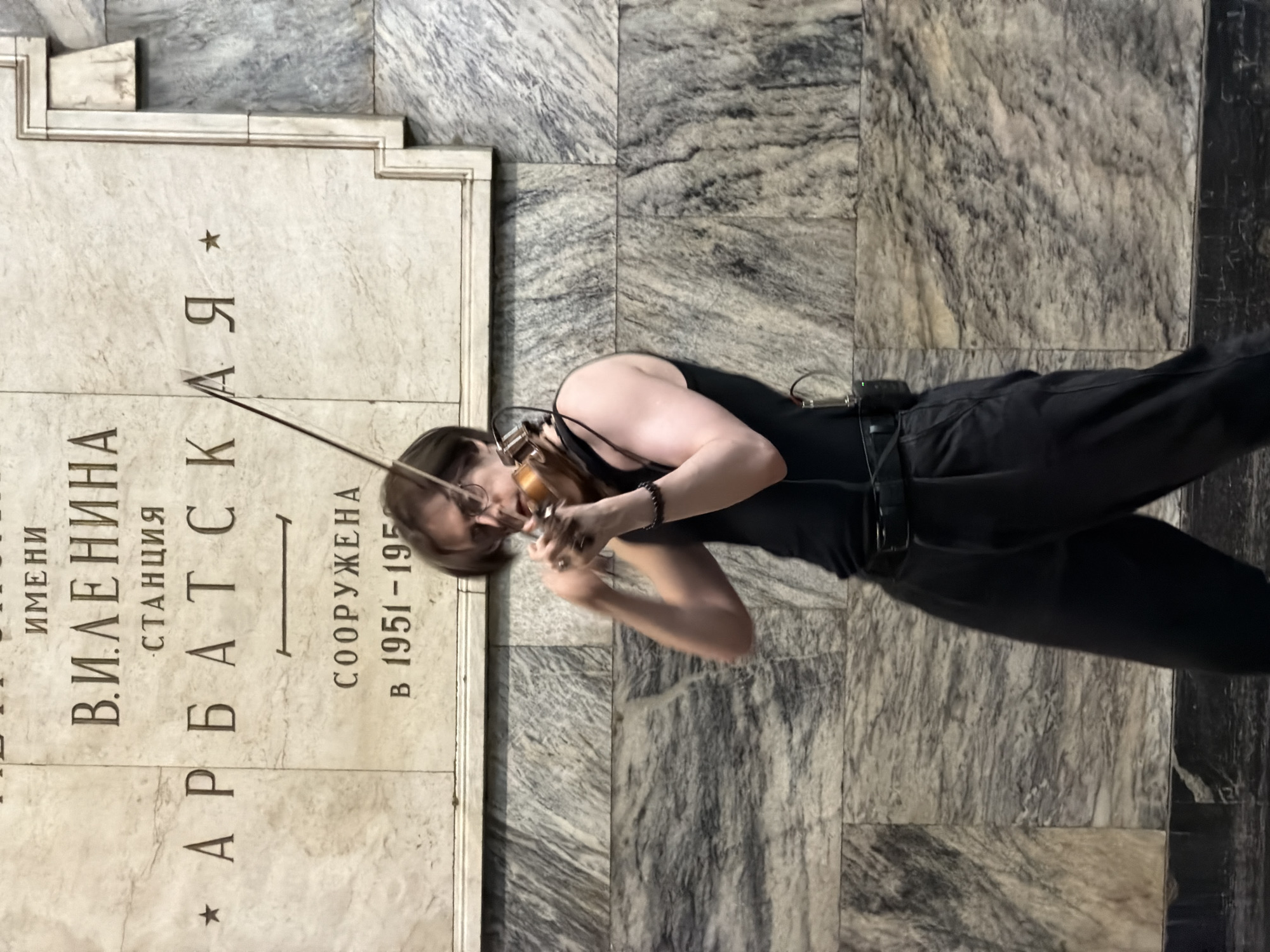 A musician, presumably a female, is playing the violin next to a marble inscription at the Arbatskaya Metro station in Moscow, Russia. The inscription is a plaque with the name of the station and construction years (1951-1953). The musician is wearing a black tank top and black trousers and playing a violin. There is a cable and microphone attached to her body, suggesting that she is recording or amplifying her music. The location is a section of the Metro station that is clad in marble.