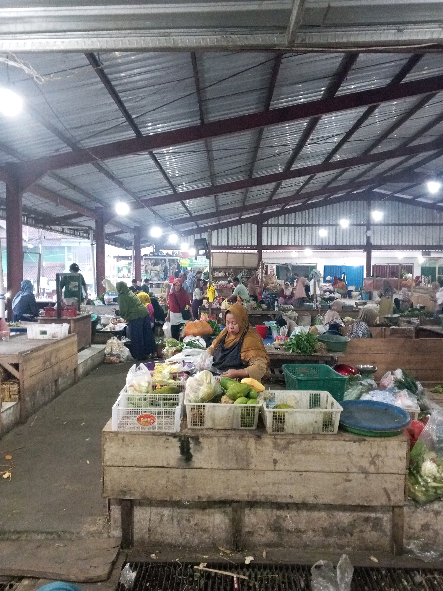 The image depicts an indoor marketplace in Gentan, Indonesia, characterized by a corrugated metal roof supported by metal beams. The space is filled with numerous stalls where vendors, mostly women wearing hijabs, are selling produce. The foreground features a stall with a wooden base laden with baskets of fruits and vegetables, including papayas, limes, and leafy greens. A woman in an orange hijab and brown apron is actively engaged at this stall. Other stalls are visible further back, displaying a variety of goods, and a diverse group of people, including vendors and customers, are interacting within the market. The ground appears to be concrete, and there is a drain grate in the lower foreground. The overall impression is a bustling local market environment.