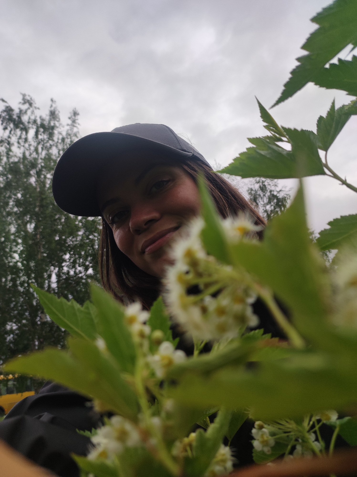 A young woman is posing for a photograph, partly obscured by a flowering shrub with white flowers and green leaves. She is wearing a dark-colored baseball cap. The background features overcast skies and blurred trees, suggesting an outdoor setting. The image was taken in the city of Chaikovsky, Russia.