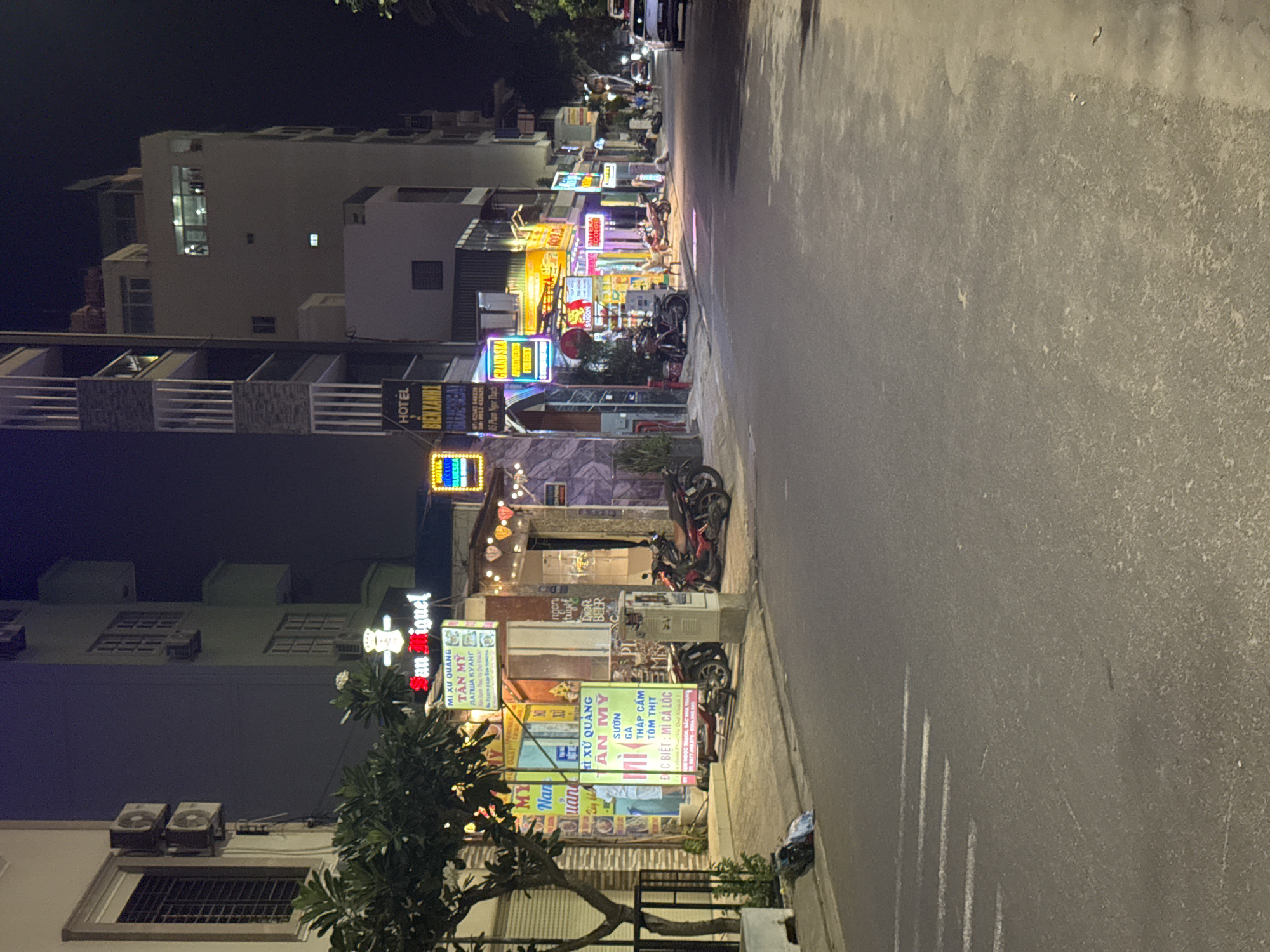 This is a detailed nighttime street scene in Bac Nha Trang Ward, Vietnam, characterized by numerous illuminated commercial signs and parked motorbikes.
On the left side of the street, a light-colored building with a window and two external air conditioning units marks the foreground. Below it, a small tree with green leaves stands beside a black metal railing and a white planter. A blue plastic bag, possibly containing trash, rests on the sidewalk. Adjacent to this, businesses are brightly lit by neon signs. Prominently displayed is a "MI XU QUANG TAN MY" sign, a Vietnamese noodle restaurant, with additional green and white signs detailing menu items like "SƯƠN GA THẬP CẨM TÔM THỊT" and "ĐẶC BIỆT: MÌ CÁ LÓC," along with phone numbers (0777.677.177 and 0904.400.303). A "San Miguel" beer sign is also visible. Further along, a business features decorative string lights resembling lanterns. Several motorbikes are parked on the sidewalk in front of these establishments. A white utility box stands on the sidewalk.
The right side of the street features a modern, multi-story building with grey and white facade elements. Below it, a black sign identifies "HOTEL BIEN XANH 1" (Blue Ocean Hotel 1), providing contact numbers (02583 540226, 0912 432525) and an address (85 Phạm Ngọc Thạch). Next to it, a bright red and blue neon sign advertises "GRAND SEA APARTMENTS FOR RENT." Further down the street, additional businesses exhibit various colorful, illuminated signs, including "SAIGON" and "ATTENRA COPP." More motorbikes are parked along this sidewalk. A few indistinct human figures are visible walking further down the sidewalk on the right.
The asphalt road, which appears dry and dark, occupies the central and foreground portions of the image. Faint, worn white markings, possibly remnants of a pedestrian crossing, are visible on the left side of the road in the mid-ground. The street recedes into the dark background, where more illuminated buildings and distant vehicle lights are discernible. The sky above is dark and clear. Artificial light from signs and unseen streetlights illuminates the scene.
