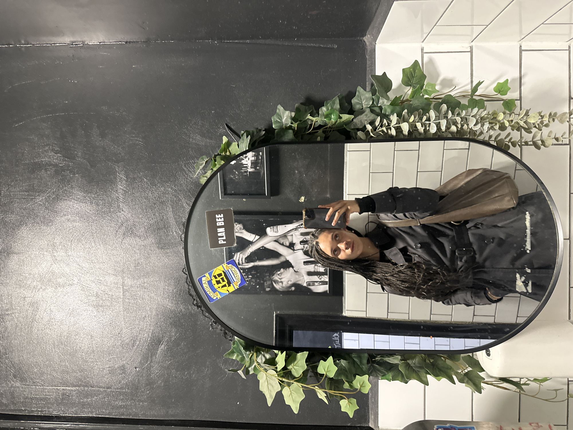 This media file features a mirror selfie taken by a person with long, dark braided hair, holding a smartphone. The person is wearing a black top or jacket and has a brown shoulder bag. Their expression is neutral to slightly smiling.
The selfie is captured in an oval-shaped mirror, which is decoratively adorned with artificial green foliage, including ivy and eucalyptus-like leaves, around its top and sides. The mirror is mounted on a textured black wall that shows some scuffs and marks. To the right, a section of clean white subway tiles is visible. This interior setting, possibly a bathroom or a decorative hallway, is located in London, United Kingdom.
Notable details in the mirror's reflection include a black and white image or poster behind the person, with the text "PLAN BEE" clearly visible. Additionally, a sticker is affixed to the mirror's surface, displaying a yellow and blue design with the text "ADMIRAL BENBOW." The overall impression is an indoor, well-lit scene, likely during the daytime.