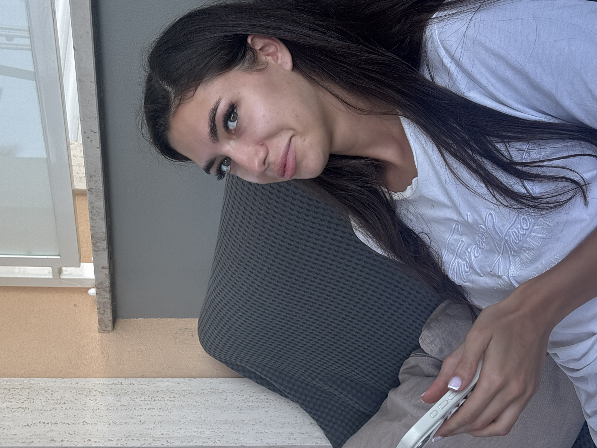 A young woman with long dark hair sits on a gray sofa, holding a white smartphone in her left hand. She wears a white t-shirt with embossed text. Her head is turned to her right, and she is looking slightly upwards and to her right. The background shows a textured cream-colored wall and a gray wall, with a glimpse of a window frame. The scene appears to be on a balcony or patio, possibly in Cupra Marittima, Italy, given the architectural style.