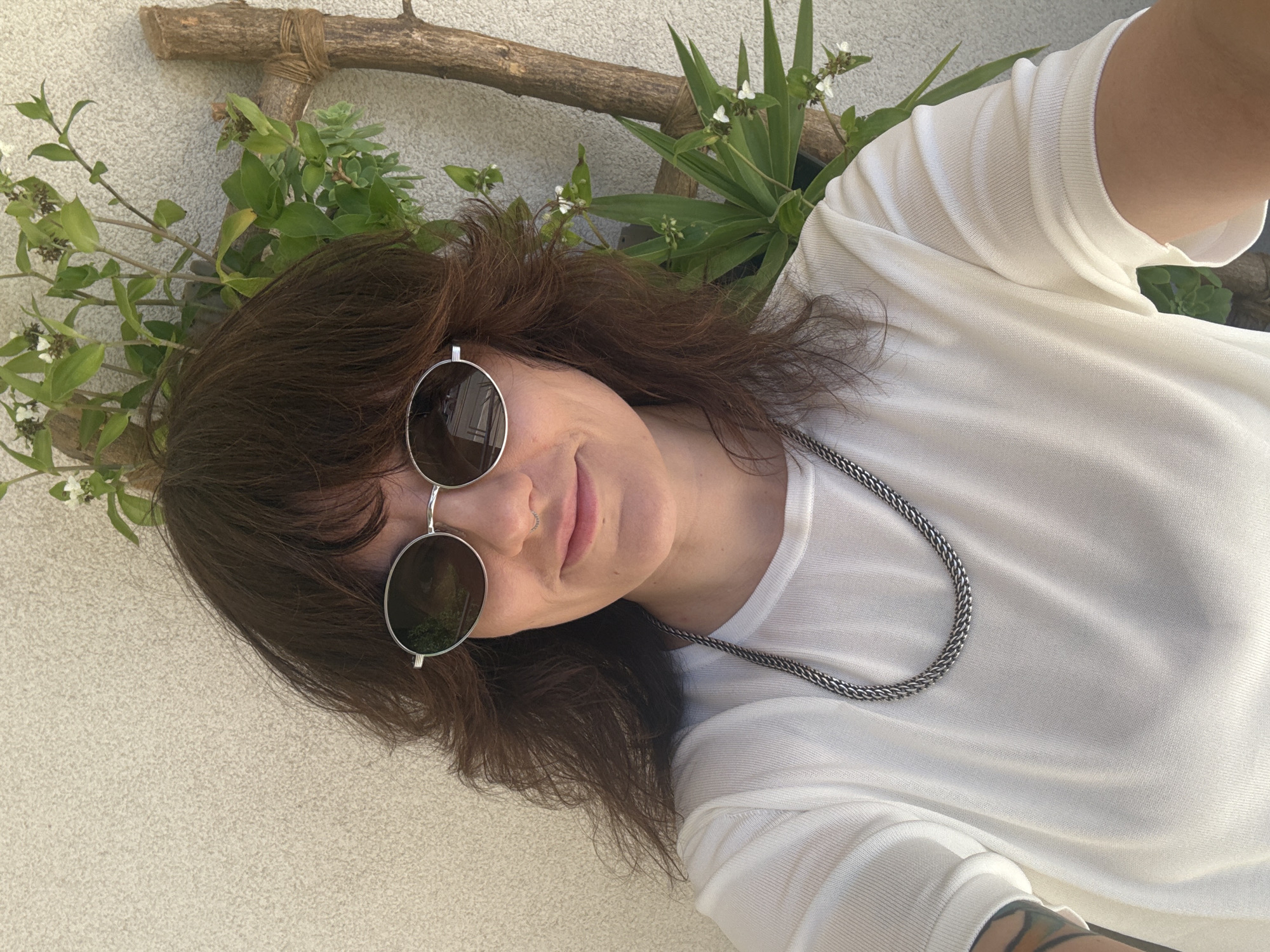A selfie of a person with brown, shoulder-length hair wearing round sunglasses and a white top. The person is wearing a silver chain necklace and has a nose ring. They are smiling slightly. In the background, there is a wall with greenery including a branch, potted plants and some foliage with small white flowers. The photo may have been taken outdoors, possibly in a garden or patio area. There is no information to confidently determine the location of the photo in the city of Budva, Montenegro.