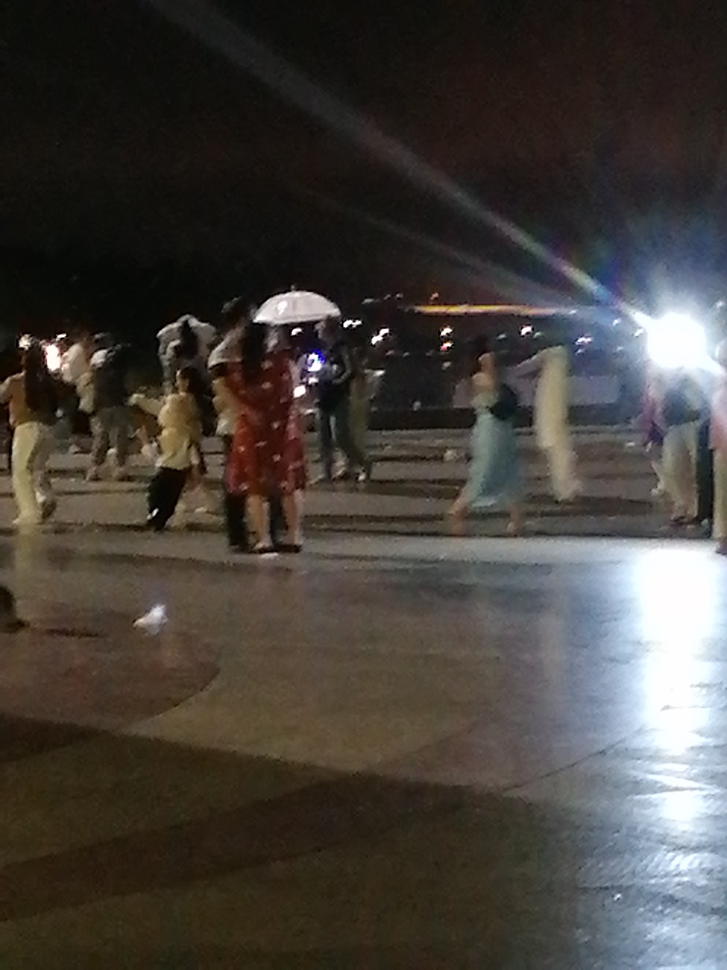 The image captures a nighttime scene in Da Lat, Vietnam, likely in an outdoor public space with a paved ground surface that appears wet, reflecting the artificial lights. A group of people are present, some appearing to be dancing or engaged in social activity. In the center, two individuals stand close together; one is wearing a red and white patterned dress, and the other, whose face is obscured, is wearing a dark top and lighter pants. To the left of this pair, a person in a yellow top and dark pants is captured mid-stride, possibly dancing. An umbrella, held by an unseen person, is visible above the crowd, suggesting it might be raining or has recently rained. Other individuals are scattered throughout the frame, some in motion and others standing. The background is dark, with some distant lights hinting at a city environment. The overall atmosphere is lively and social, typical of a public gathering at night.