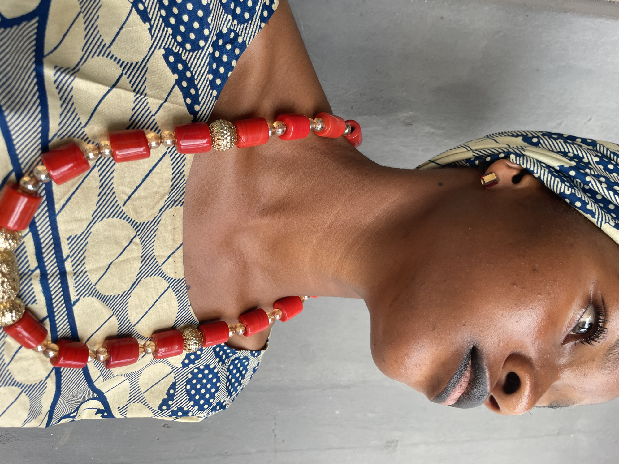 A close-up shot shows a dark-skinned woman wearing traditional Nigerian attire, likely in Badagry, Nigeria. She wears a patterned head wrap and a top featuring cream and blue designs. A red and gold beaded necklace adorns her neck. The woman has dark lipstick and an earring. The background is a plain grey surface.