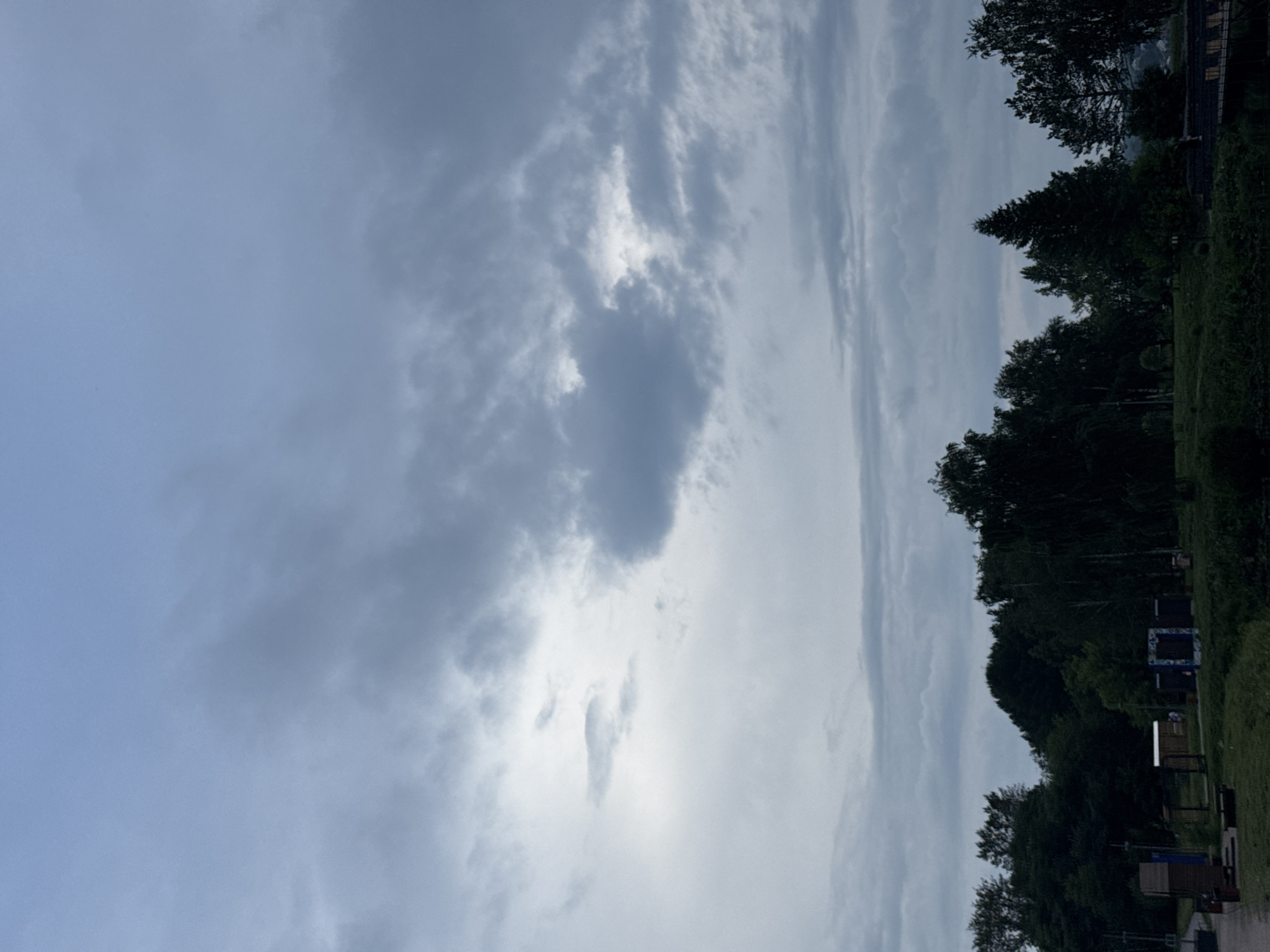 The image captures a cloudy sky over a natural landscape that appears to be located in or near Lysva, Russia. The sky is filled with a variety of gray and white clouds, with some patches of lighter sky visible. Below the sky is a dense line of dark green trees, forming a forest that stretches across the horizon. In the foreground, there are some structures, possibly part of a park or recreational area, including what looks like a wooden shed, a bench, and some colorful decorative frames or art installations. A body of water, likely a river or pond, is present in the lower part of the image, with some darker vegetation along its banks. The overall atmosphere of the scene is subdued and natural, with no people or animals visible.